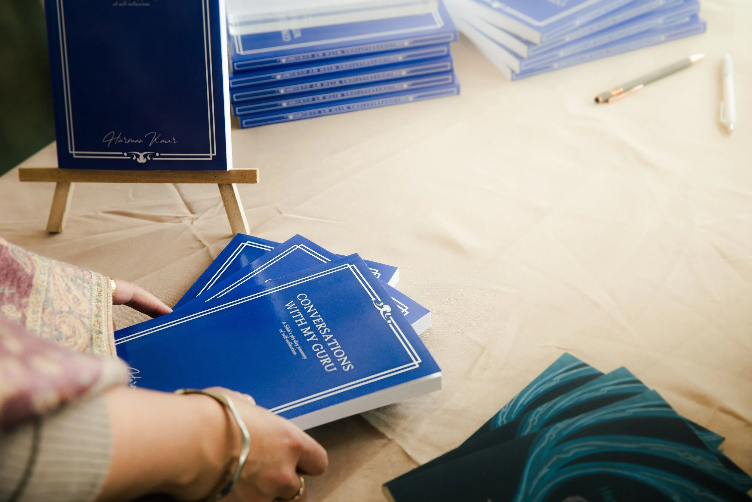 Person handling blue booklets titled 'Conversations with My Guru' at a table with stacks of the same booklets and writing utensils.