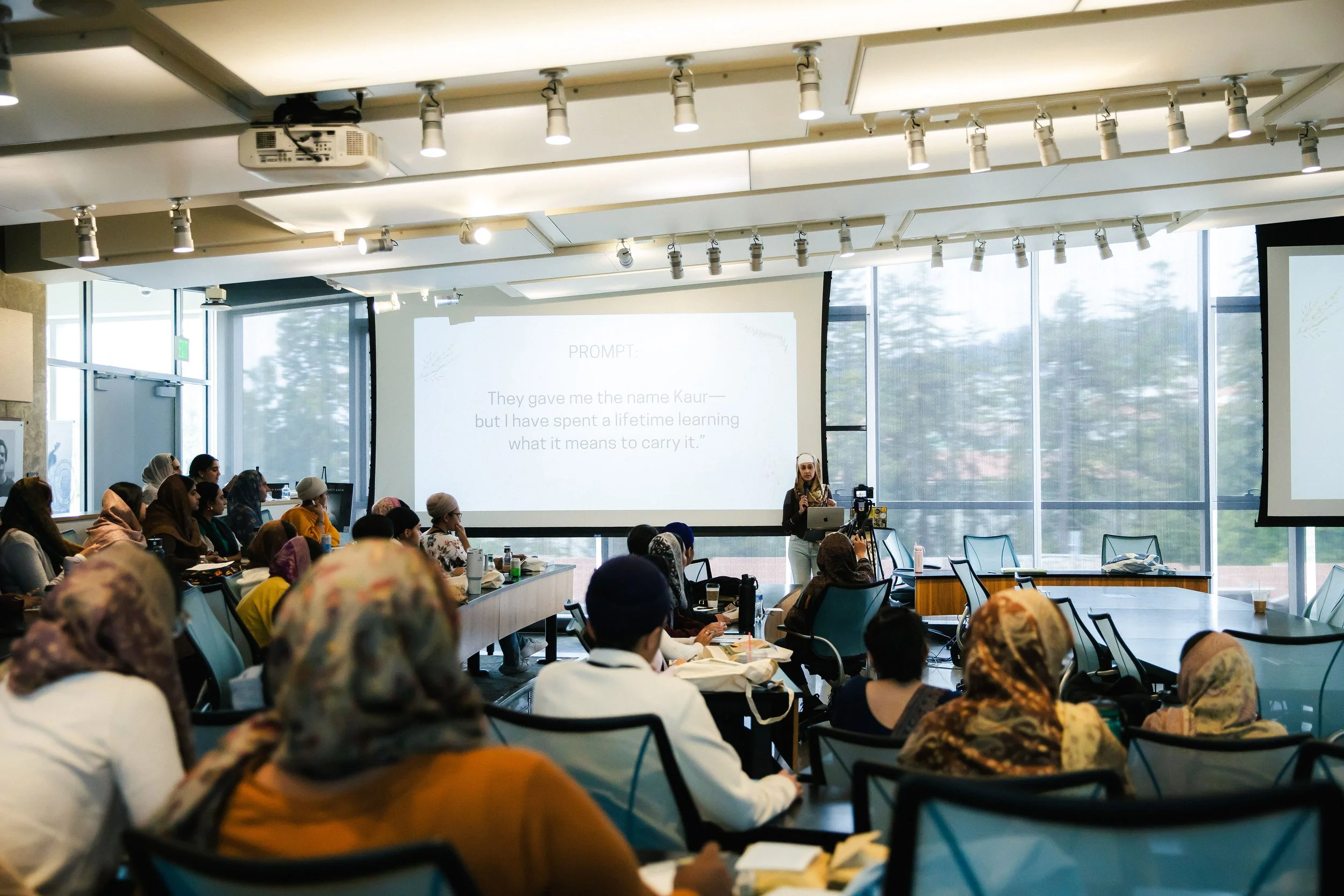 A woman giving a presentation in a conference room filled with diverse attendees, some wearing headscarves, with a large screen displaying a prompt.