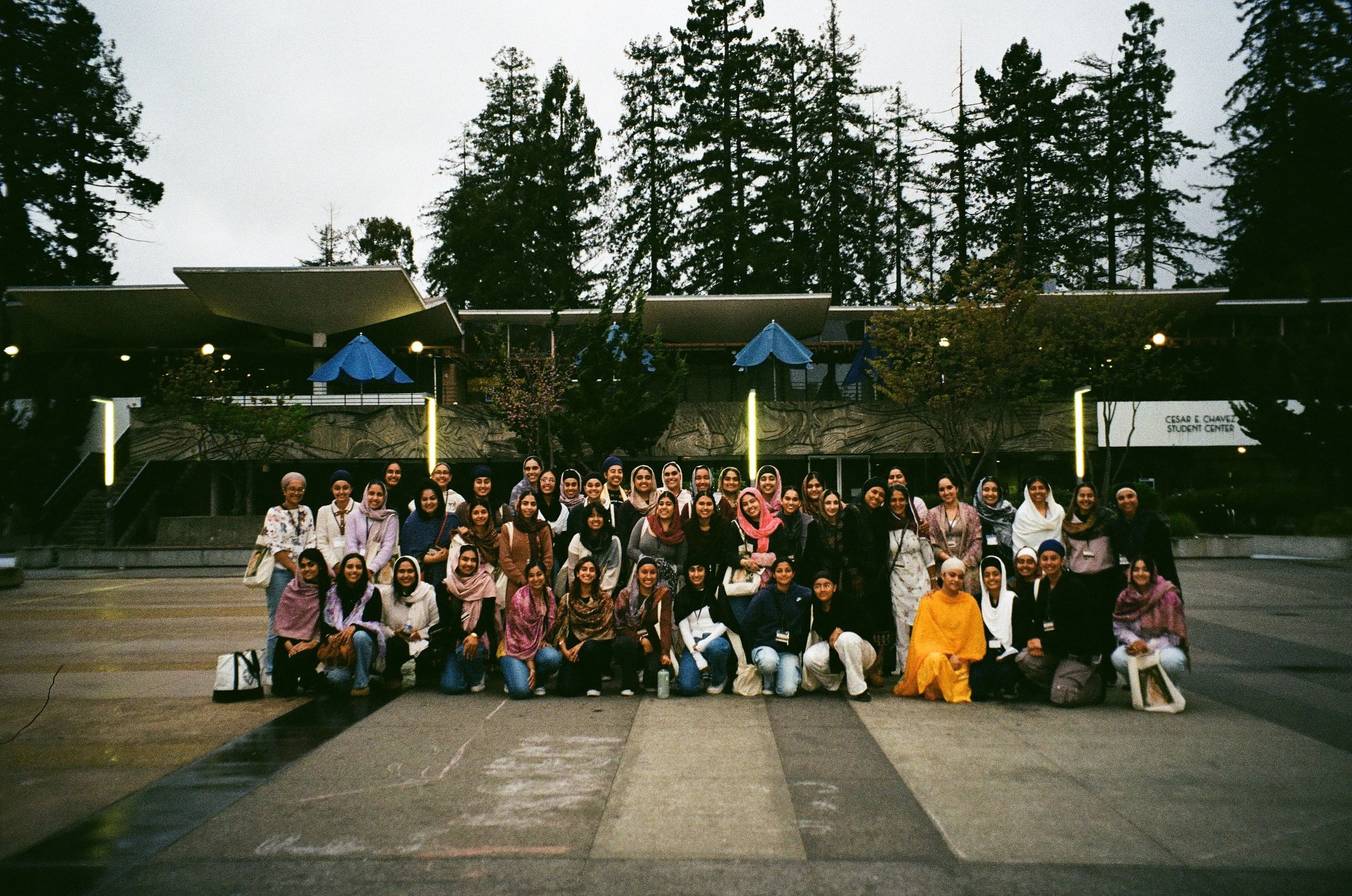 Group of diverse people posing for a photo outdoors in front of a building with tall trees behind.