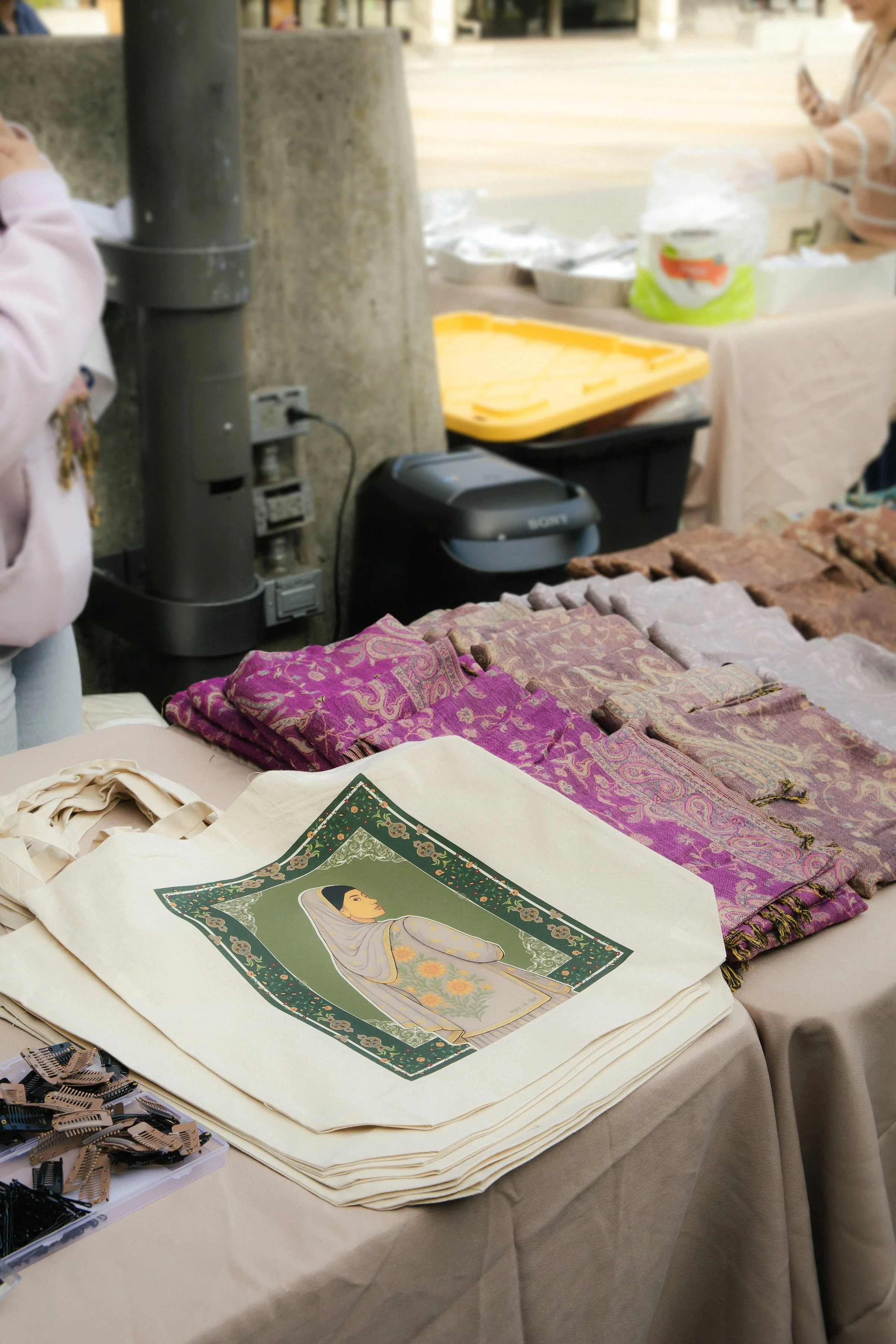 A table at a market stall displaying tote bags with a woman's portrait and patterned scarves, along with hair clips, outdoors.