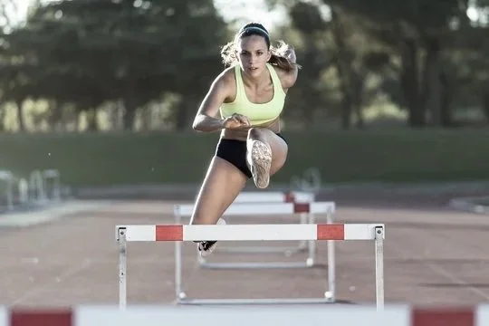 A female athlete in a yellow tank top and black shorts is competing in a hurdle race on an outdoor track.