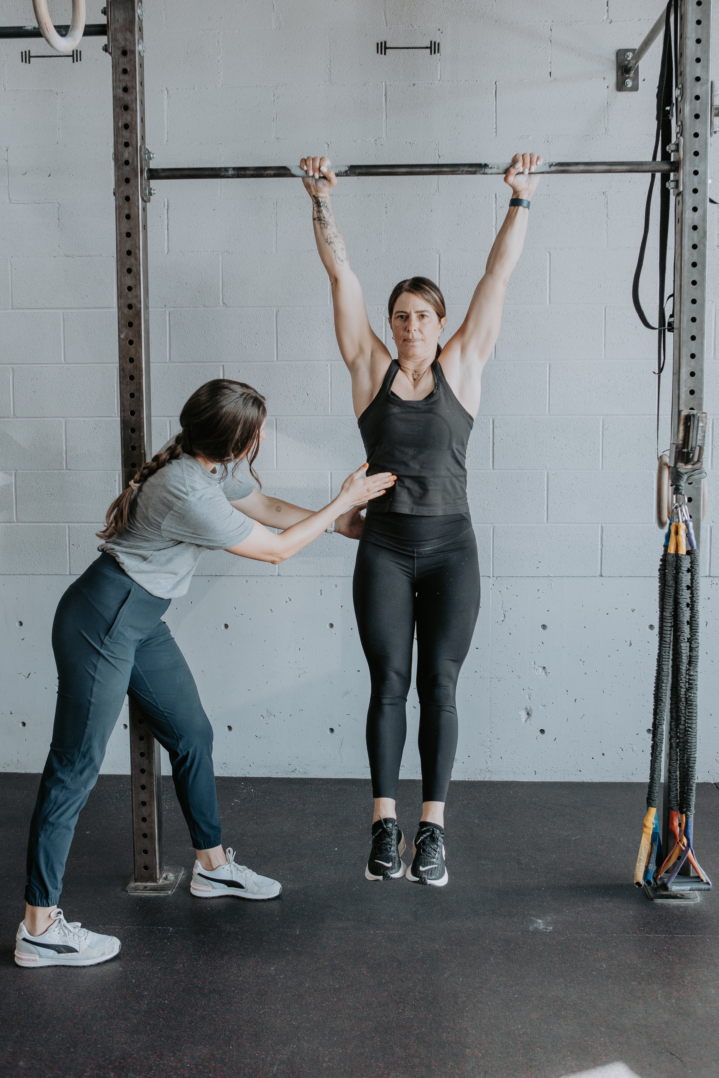A woman performing a pull-up in a gym, assisted by another woman.