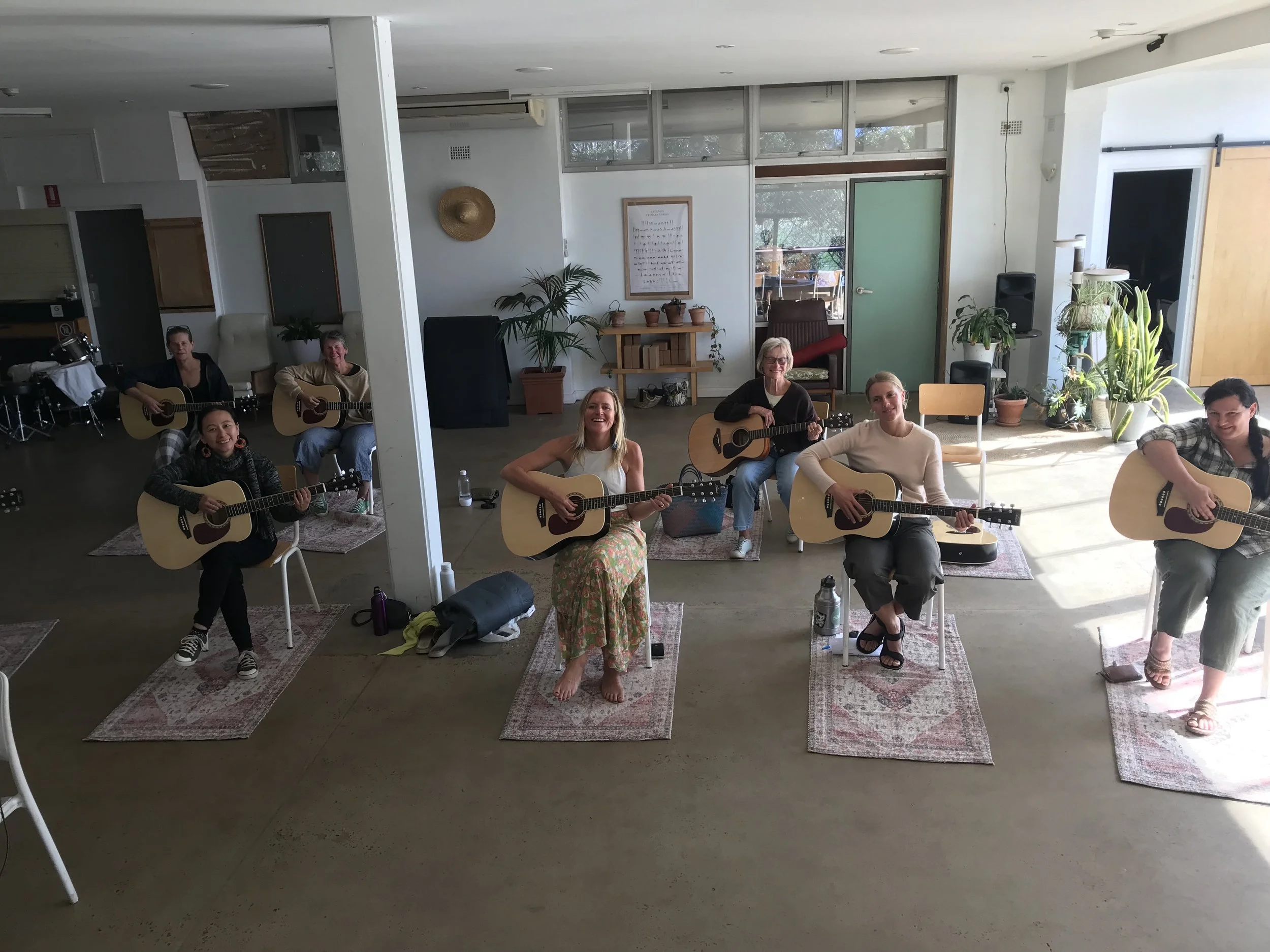 A group of eight women sitting on chairs and sitting on the floor, each playing an acoustic guitar in a bright, cozy room with potted plants and large windows.