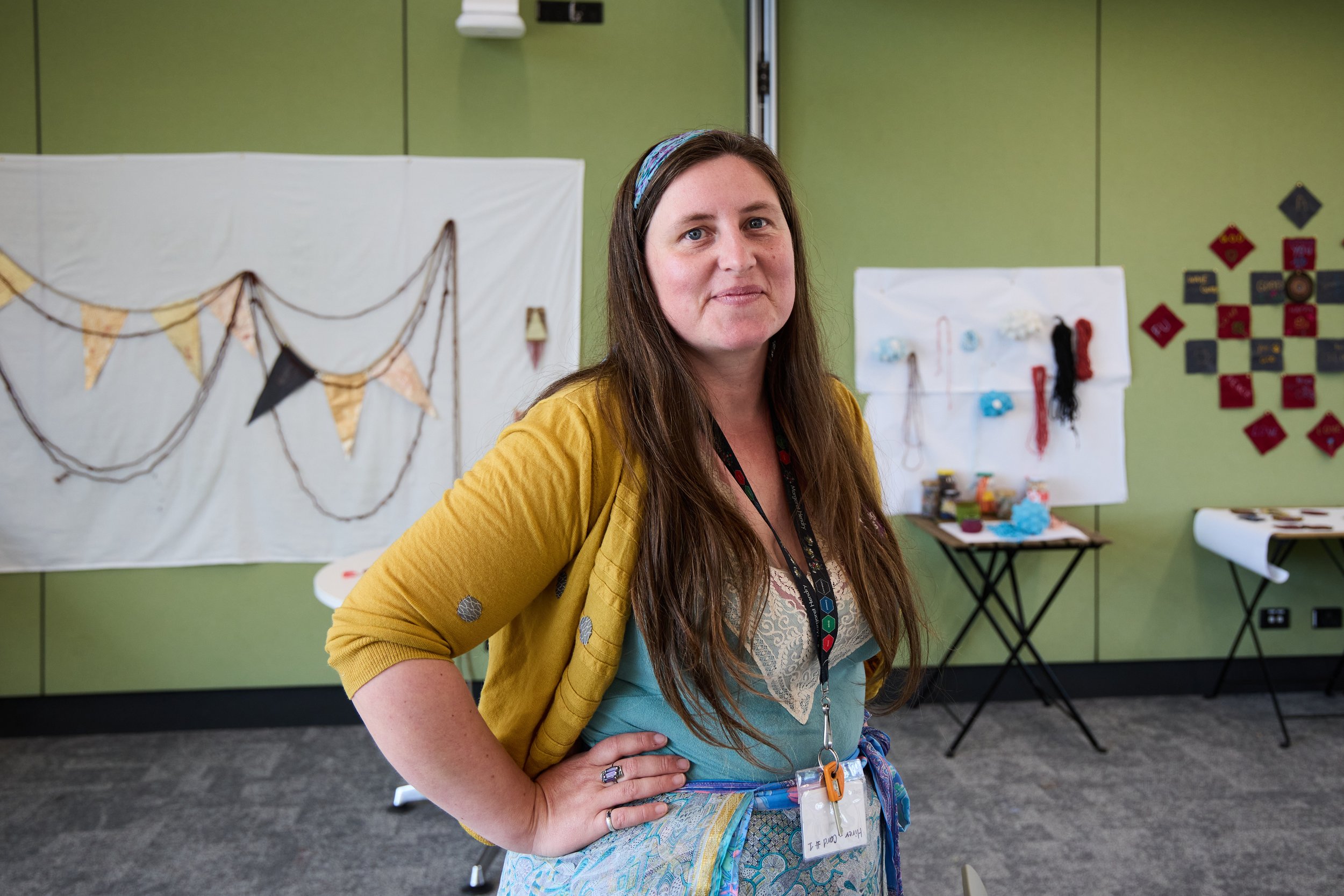 A woman with long brown hair and a headband, wearing a yellow cardigan over a teal top, stands in a room with creative decorations on the walls. She has her hand on her hip and is looking at the camera. Behind her are display boards with various craft items.