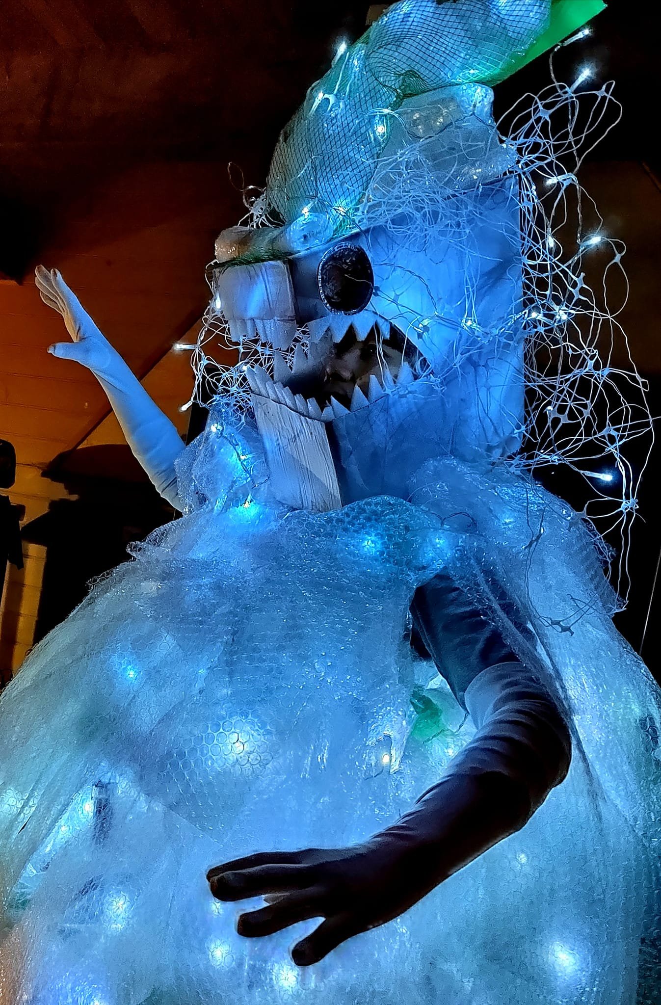 A person dressed in an elaborate costume resembling a monstrous creature with sharp teeth, a large eye, and glowing lights, surrounded by tangled LED string lights and bubble wrap, creating a spooky display.