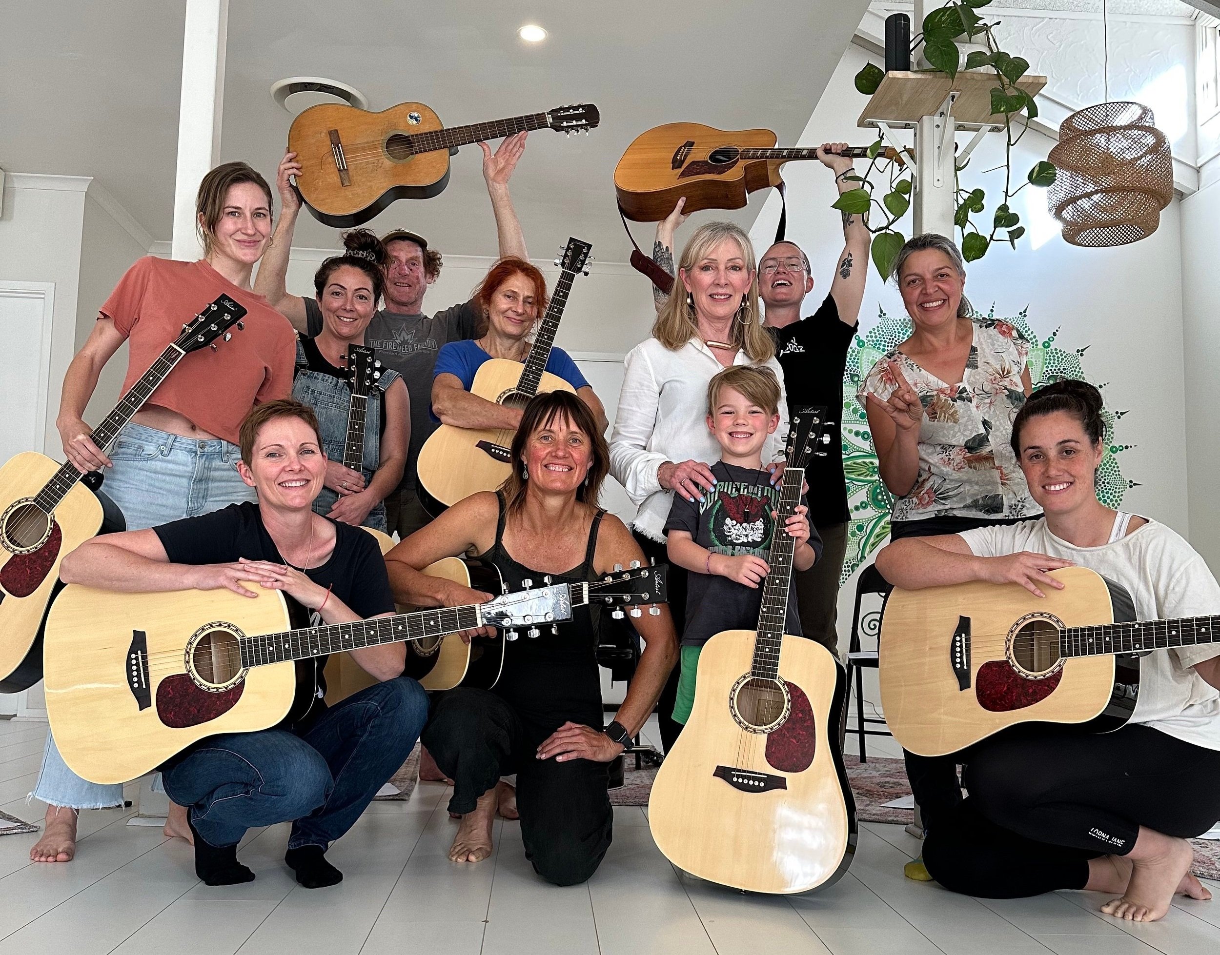 A group of eleven people, including children, posing with guitars in a well-lit indoor space. Some are standing, others kneeling or sitting, holding guitars, with a potted plant and artwork in the background.