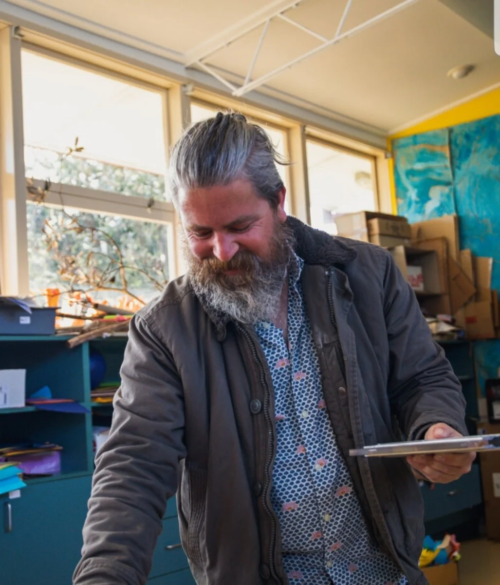 A man with gray hair and a beard smiling and looking down in a room with large windows, colorful abstract wall art, and shelves filled with boxes and art supplies.