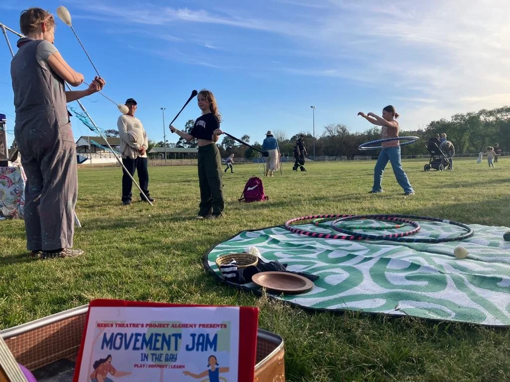 Children and adults at a park participating in a hula hoop activity, with hula hoops on the ground and a sign for a movement jam event in the foreground.