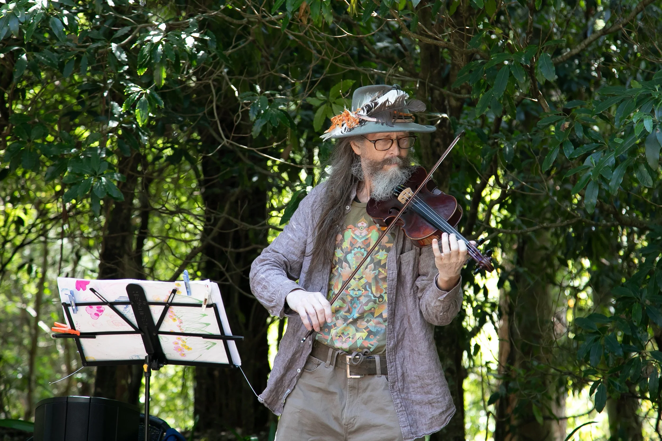 A man with long hair and a beard, wearing a decorated hat with feathers, glasses, a colorful shirt, and a gray jacket, playing a violin outdoors in a forested area.