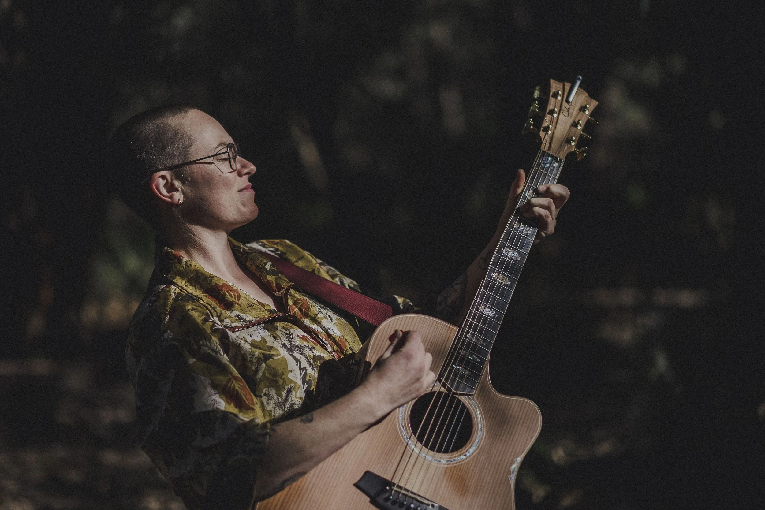 A woman with short hair, glasses, and tattoos playing an acoustic guitar outdoors at night.
