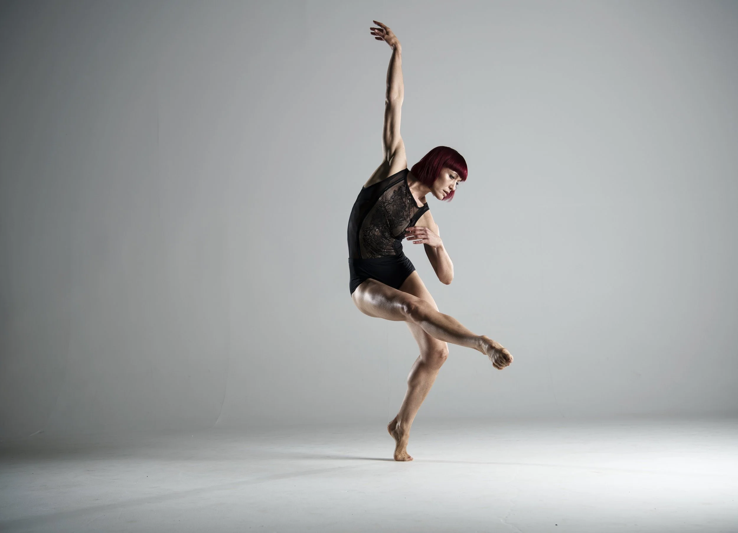 A woman with red hair wearing a black lace top and shorts dancing against a plain gray background.