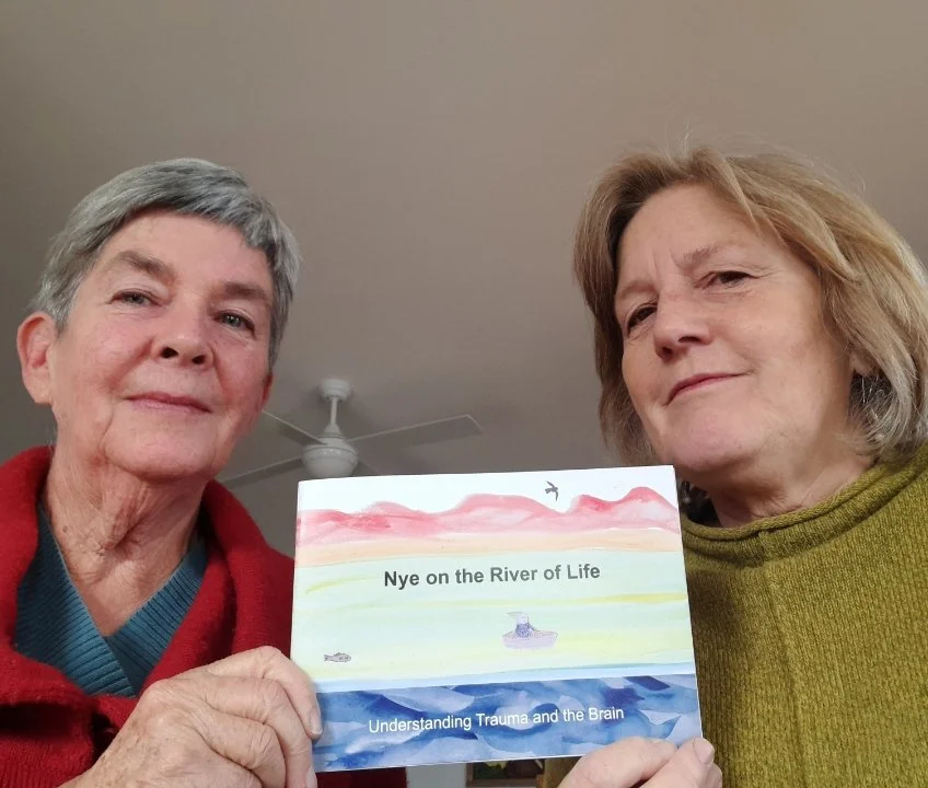 Two women holding a book titled 'Nye on the River of Life,' with a colorful illustration of a river, boat, and sky. They are standing indoors with a ceiling fan visible in the background.