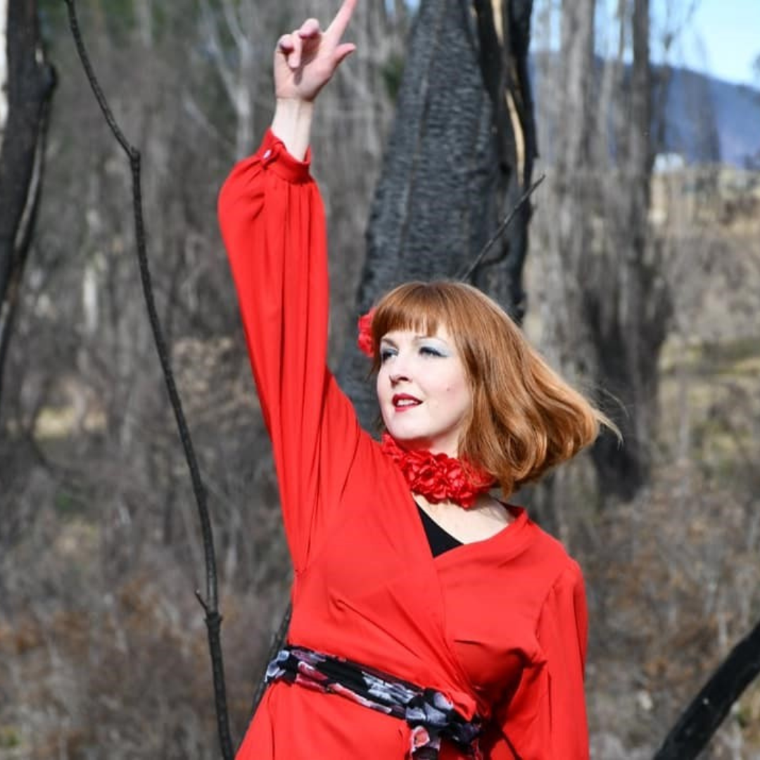 A woman with red hair in a red dress standing outdoors in a wooded area.