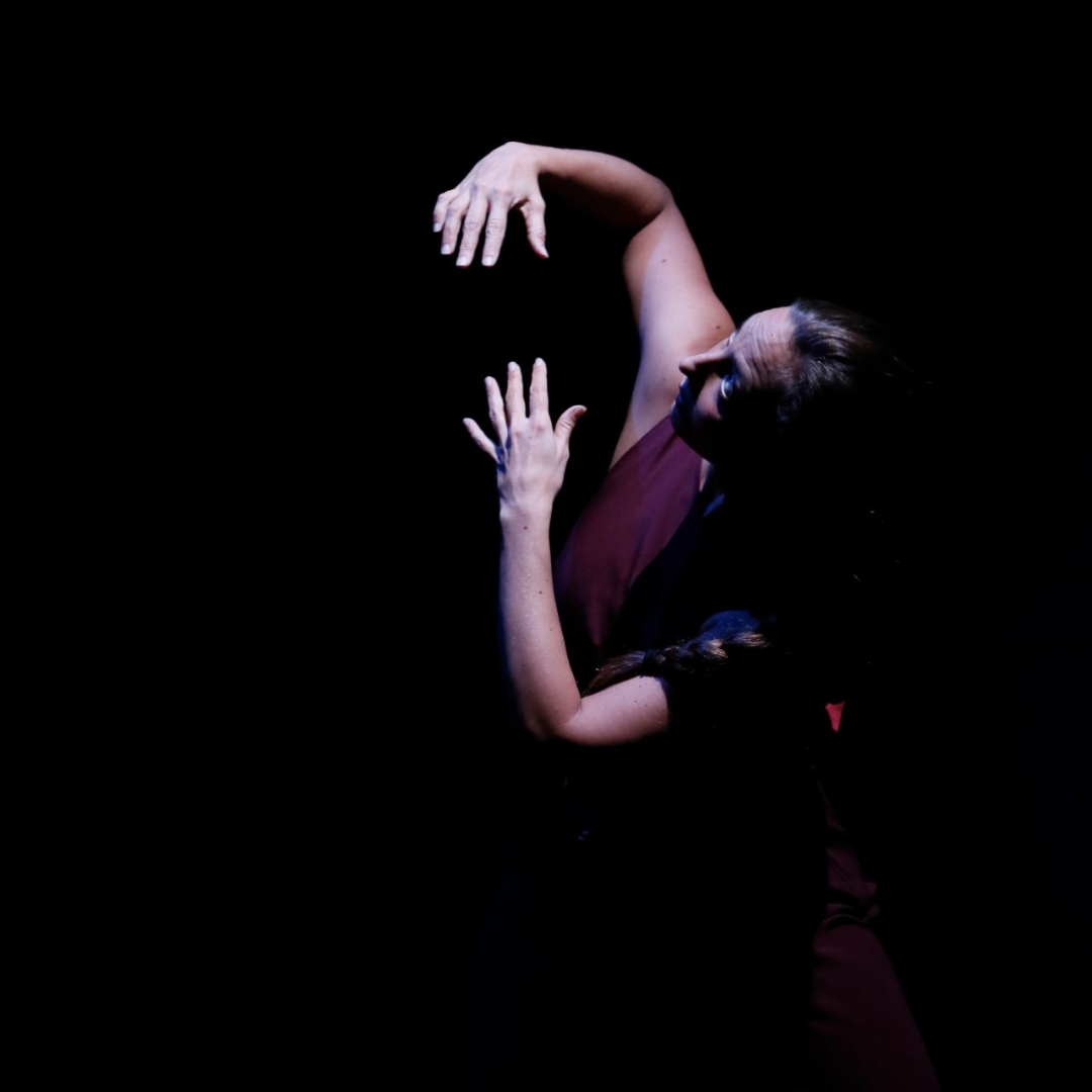 A woman performing a dance move against a dark background, with her arms raised and her head tilted back.