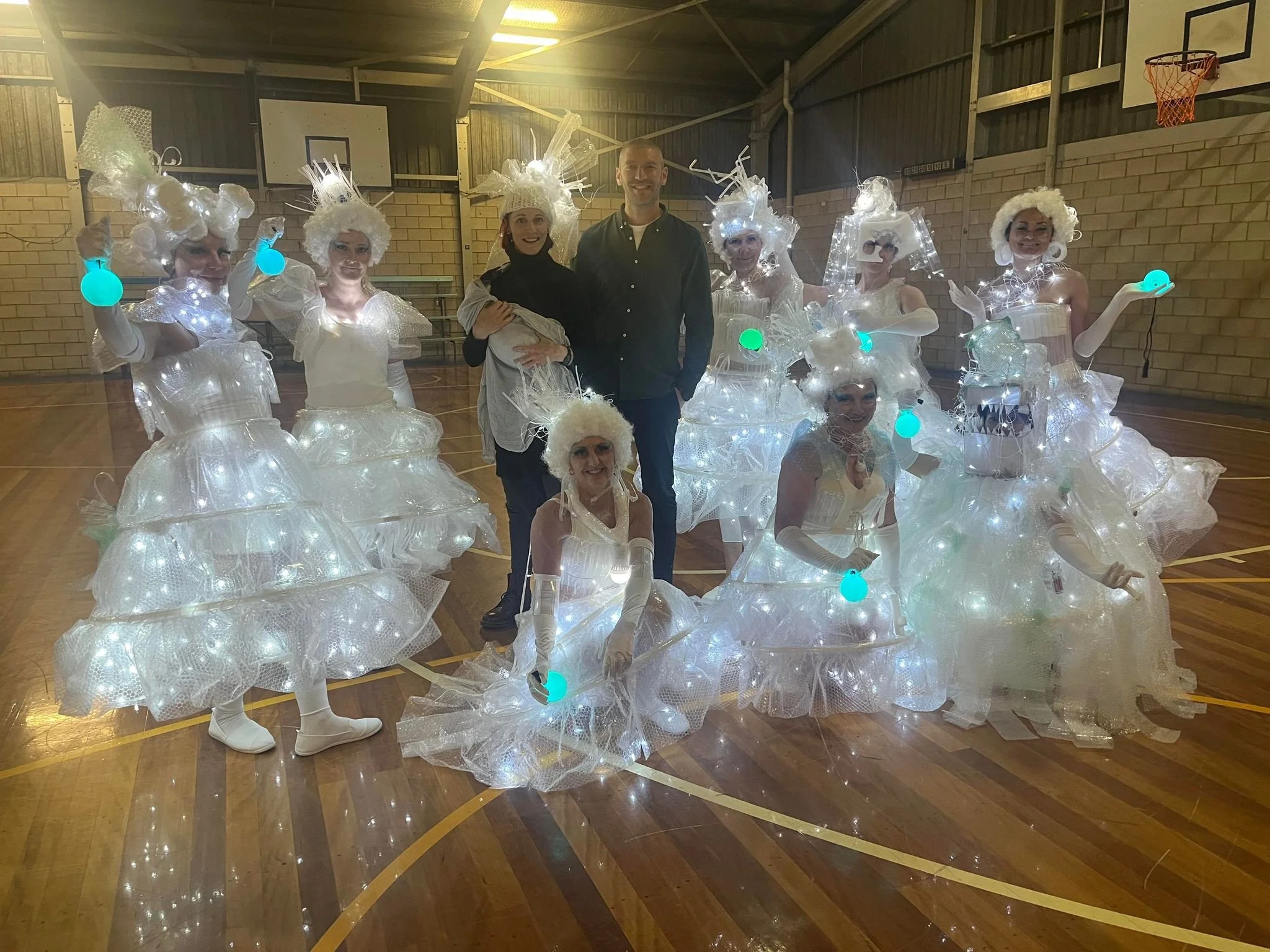 People dressed in elaborate white costumes with lights, resembling modern Christmas trees, in a gymnasium.