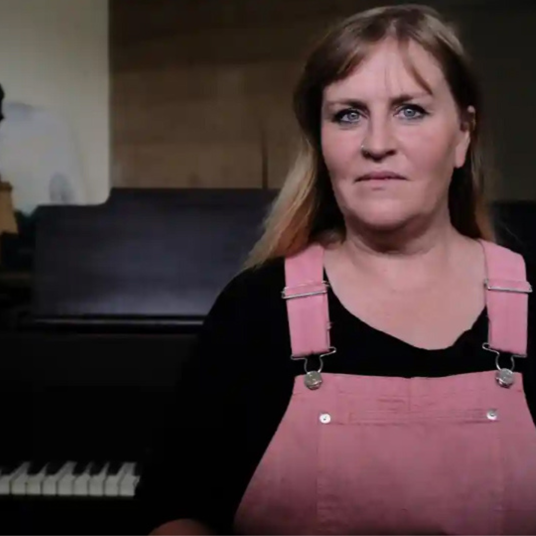 A woman with light skin, light brown hair, blue eyes, wearing a black shirt and pink overalls, standing in front of a piano in a room with wooden walls.
