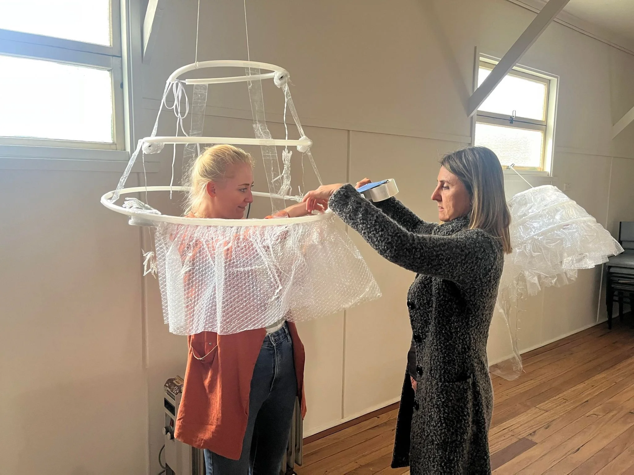 Two women inside a room with natural light, one placing bubble wrap and tape on the other, who is standing inside a large, circular bubble wrap costume with a frame.