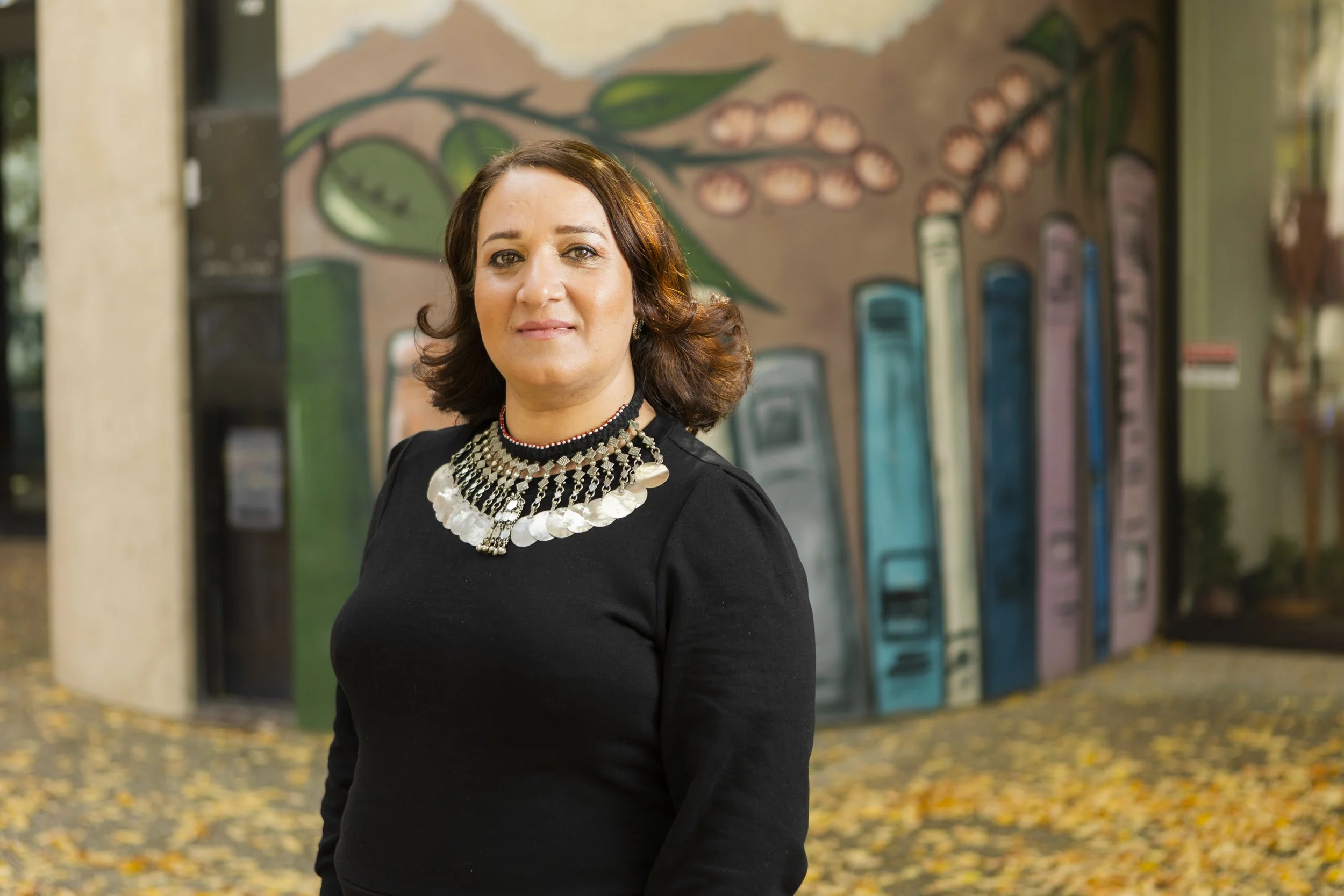 A woman with shoulder-length brown hair, wearing a black top and an elaborate silver necklace, standing outdoors in front of a colorful mural of a row of payphones, with fallen leaves on the ground.
