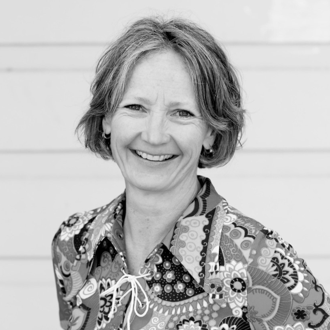 A black and white portrait of a middle-aged woman with short, wavy hair, smiling, wearing a patterned shirt with a lace-up collar.
