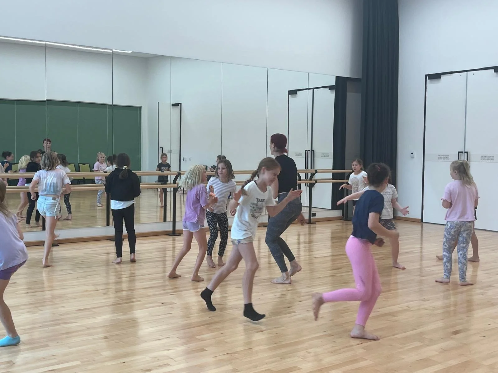 Children participating in a dance class in a studio with wooden floors, large mirrors, and ballet barres, led by an instructor.