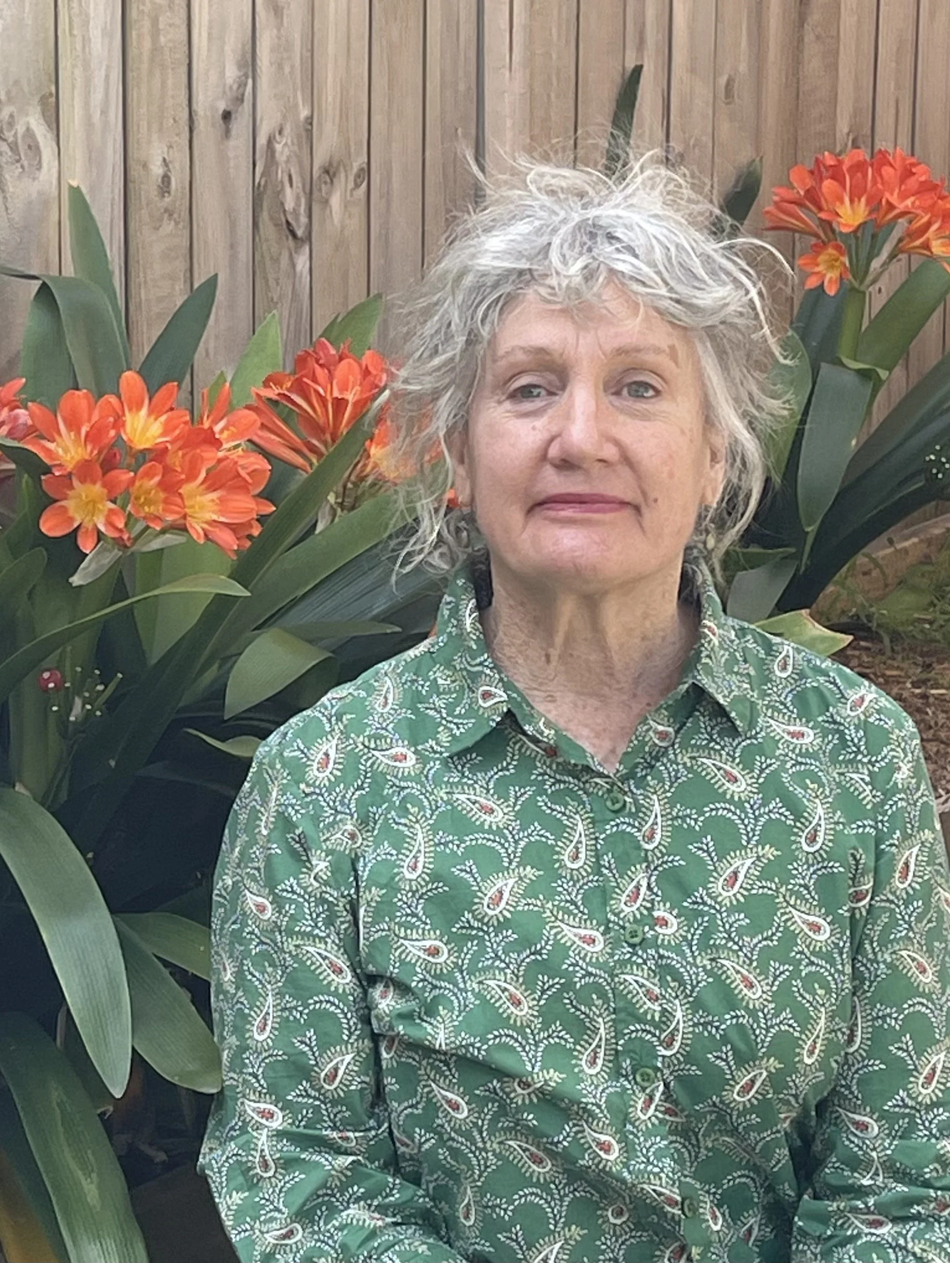 An older woman with curly gray hair sitting in front of orange flowers and a wooden fence, wearing a green patterned shirt.