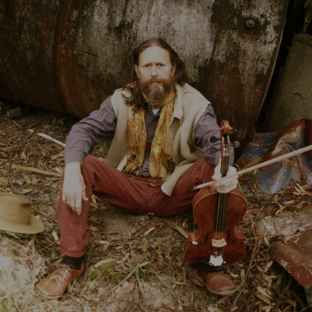 A man sitting on the ground outdoors surrounded by leaves and debris, holding a violin and bow, with a rusty metal tank in the background.