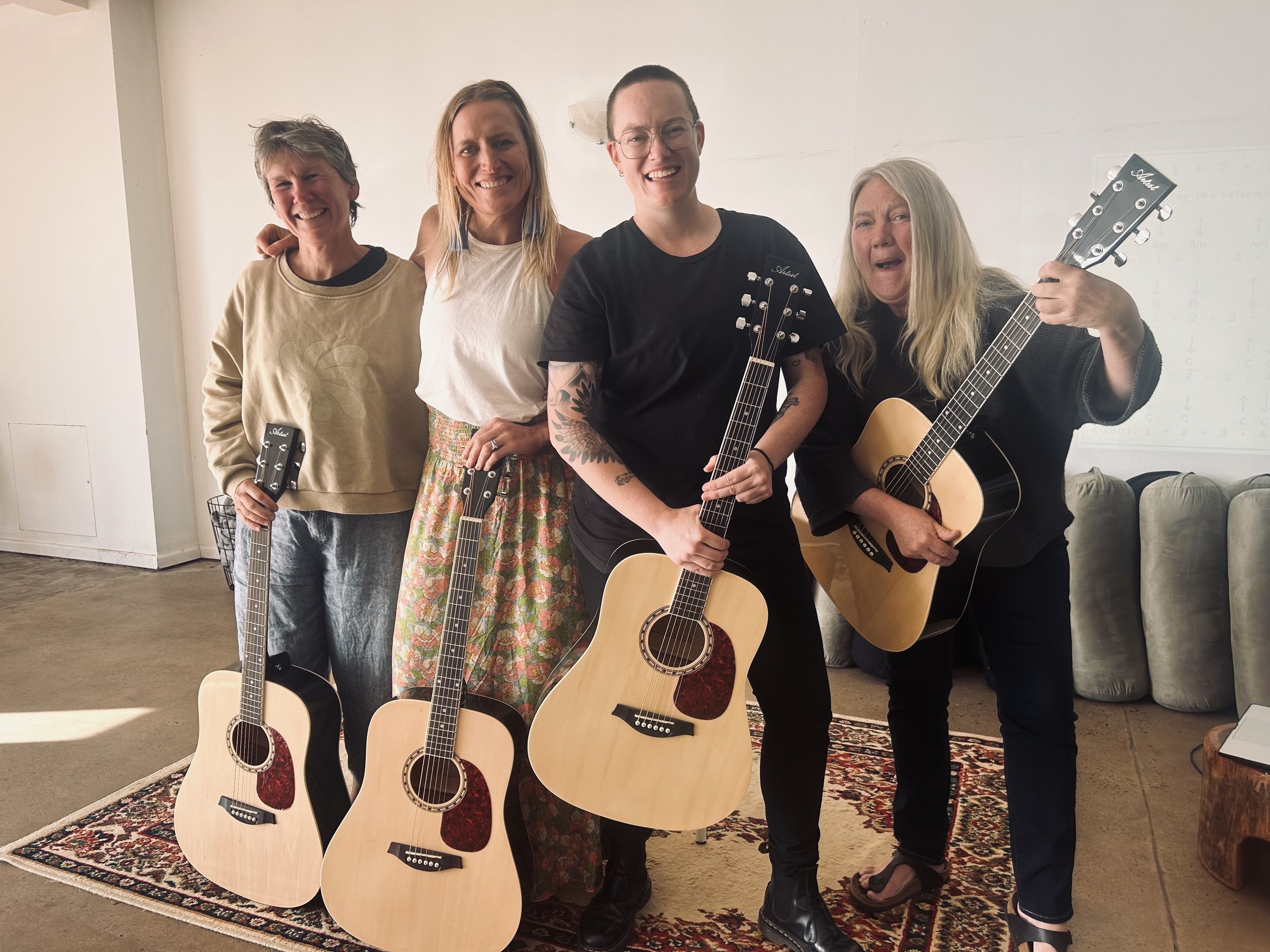 Five women posing with guitars in a room with a white wall and patterned rug.