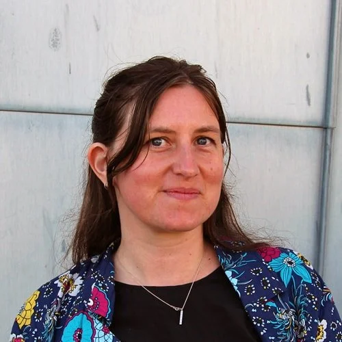 A woman with brown hair, wearing a floral jacket and a black shirt, standing in front of a gray metal wall.