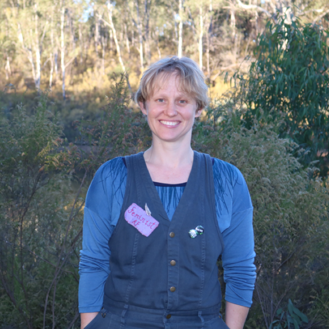 A woman with short, blonde hair smiling outdoors with trees and bushes in the background, wearing a blue long-sleeve shirt and a dark vest.