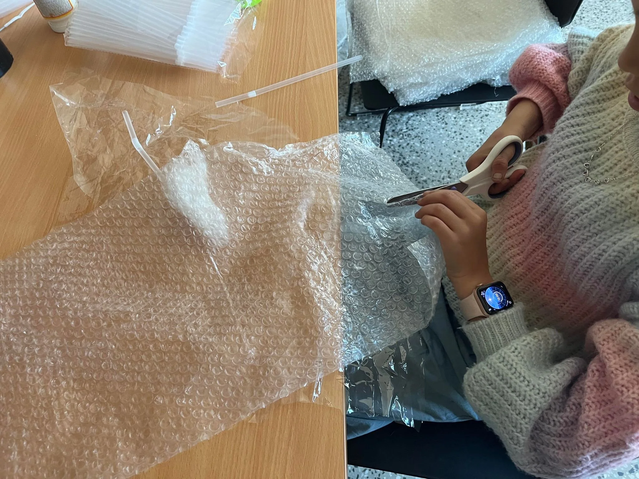 A person wearing a knitted sweater and an Apple Watch, cutting a piece of foam from a larger piece of foam wrapped in plastic bubble wrap, on a wooden table with building materials.
