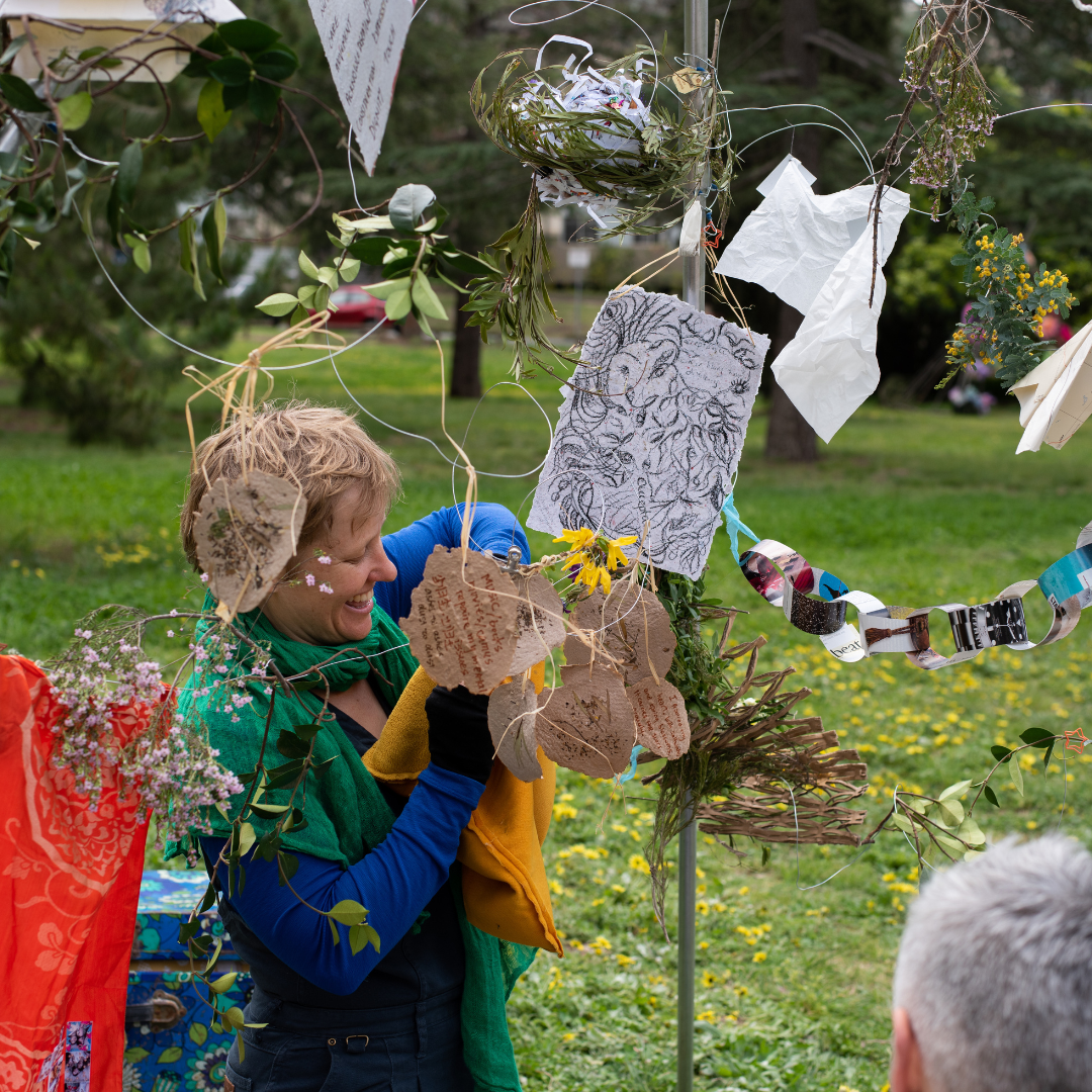 A woman smiling and decorating an outdoor installation with paper, fabric, and plant materials during a garden event.