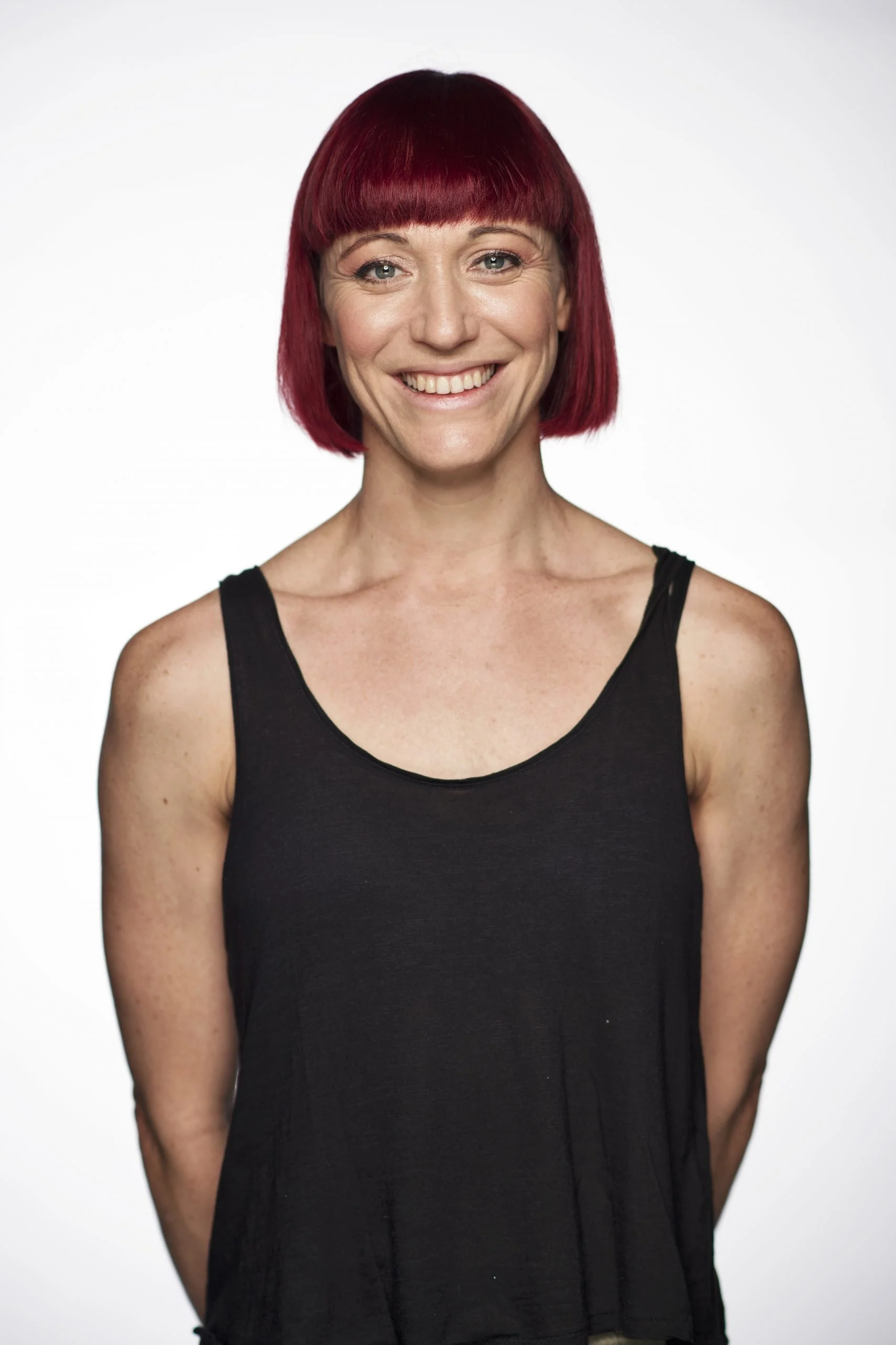 A woman with red hair and a wide smile, wearing a black sleeveless top, standing against a plain white background.