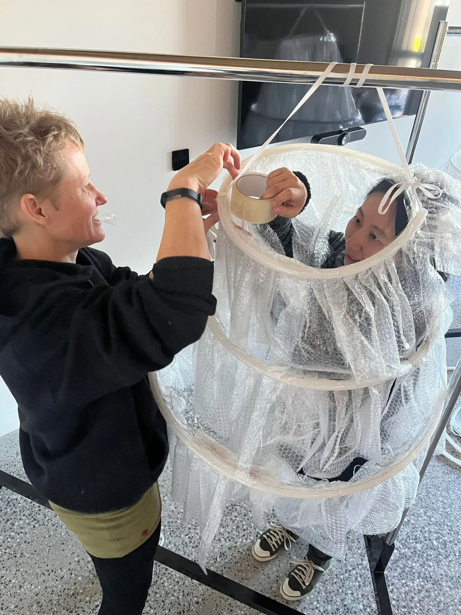 Two women assembling a bubble tent with clear panels in an indoor setting, one woman with short blonde hair smiling and the other with short black hair, wearing black and white sneakers.