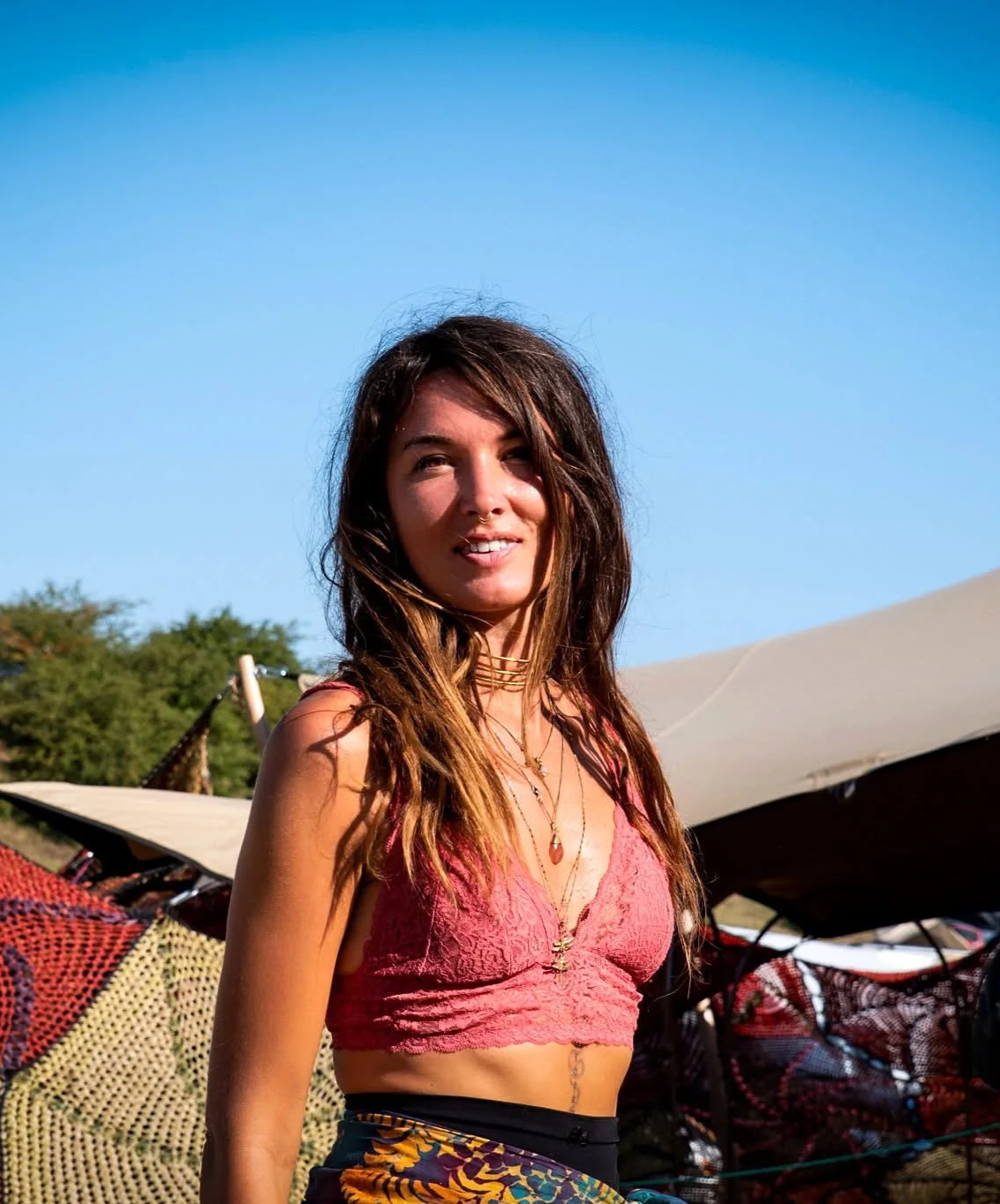 A young woman with long brown hair, wearing a pink lace crop top, standing outdoors with blue sky and tents in the background.