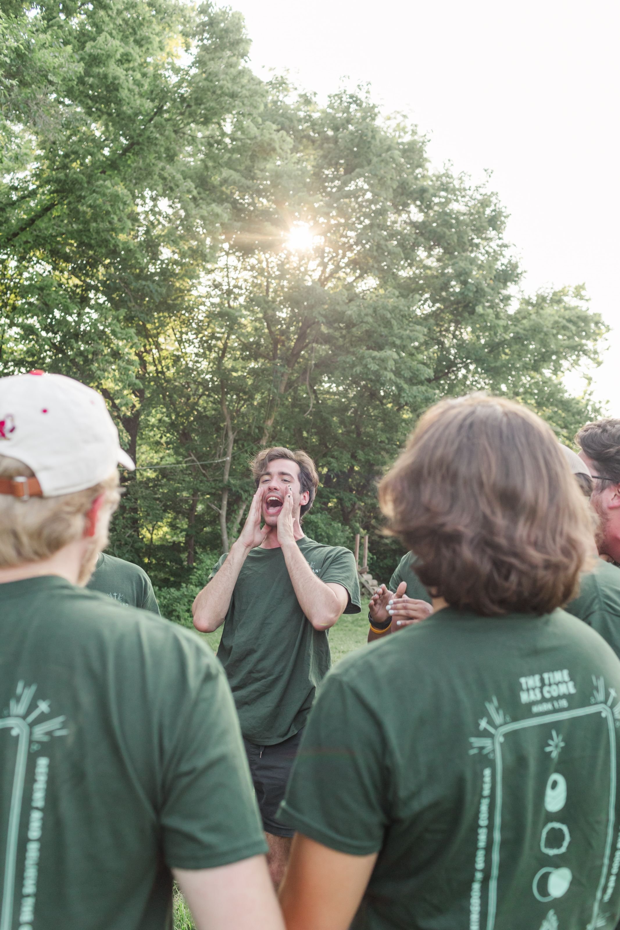 Group of Christian college students in Heartland's Gap year program gathered outdoors, one man in the center shouting with hands on his cheeks, others listening, trees and sunlight in the background.