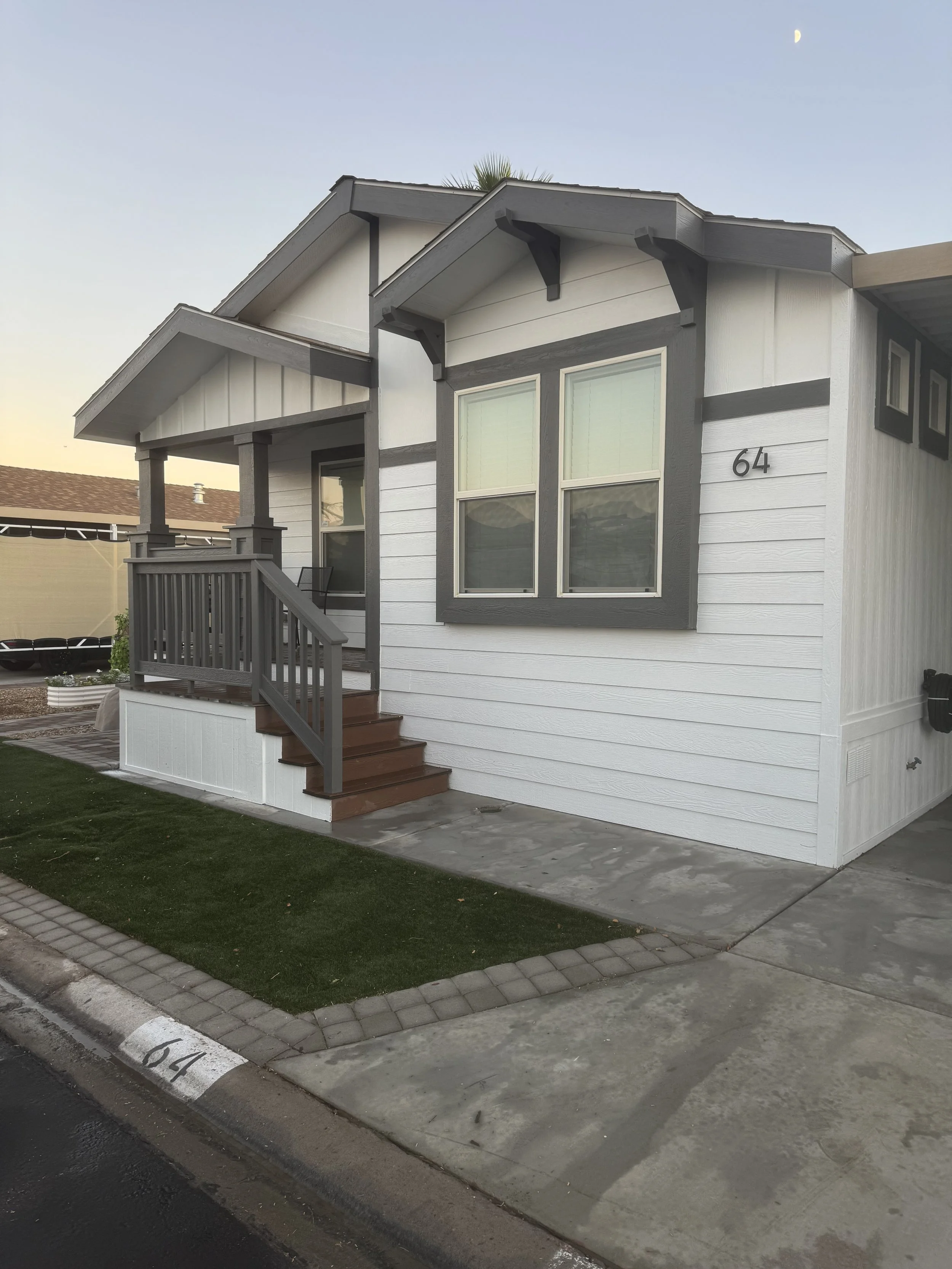 White house with gray trim, front porch, stairs, and two windows, house number 64, during daytime.