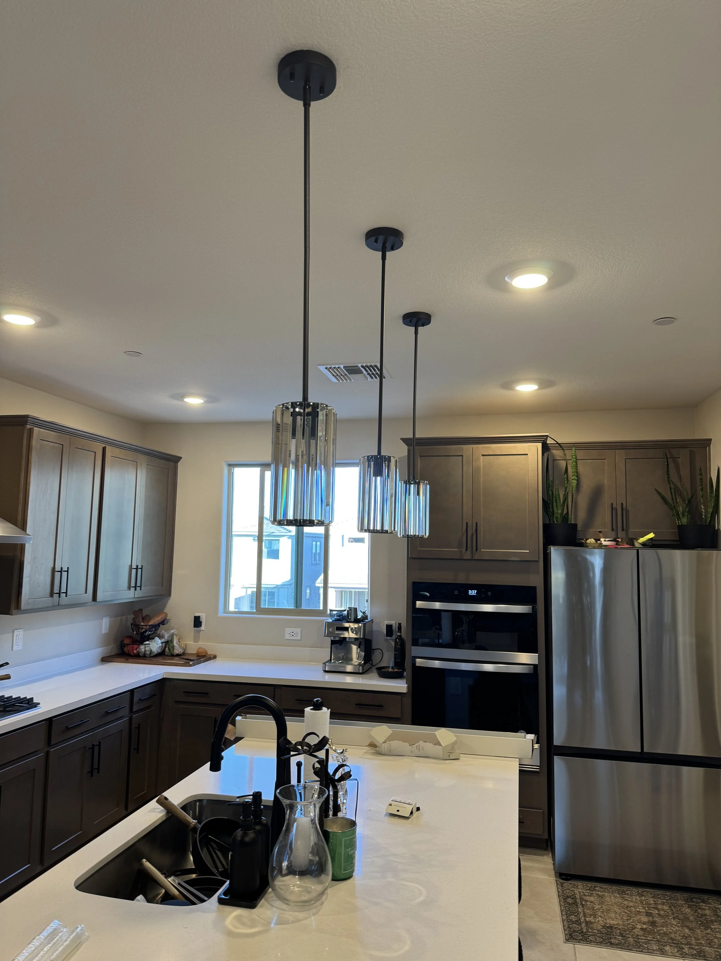 Modern kitchen with white countertops, dark brown cabinets, a stainless steel refrigerator, built-in oven, coffee maker, and decorative hanging pendant lights, with potted plants on top of the cabinets.