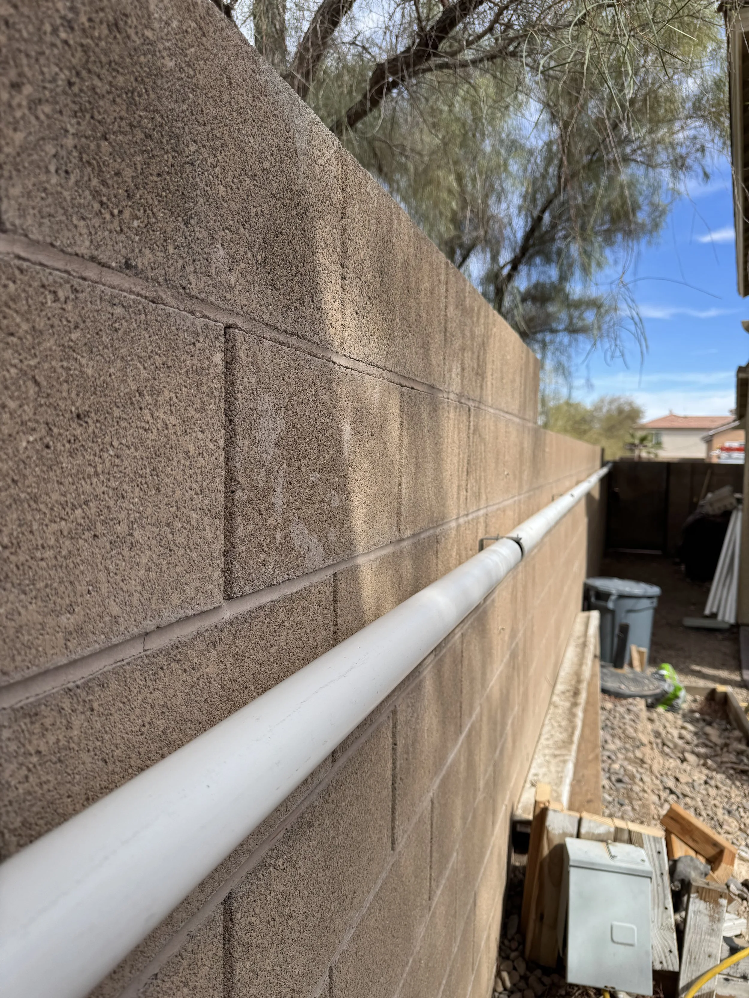 Close-up of a white pipe running along a brown brick wall outside, with construction materials and a trash bin visible in the background under a blue sky.