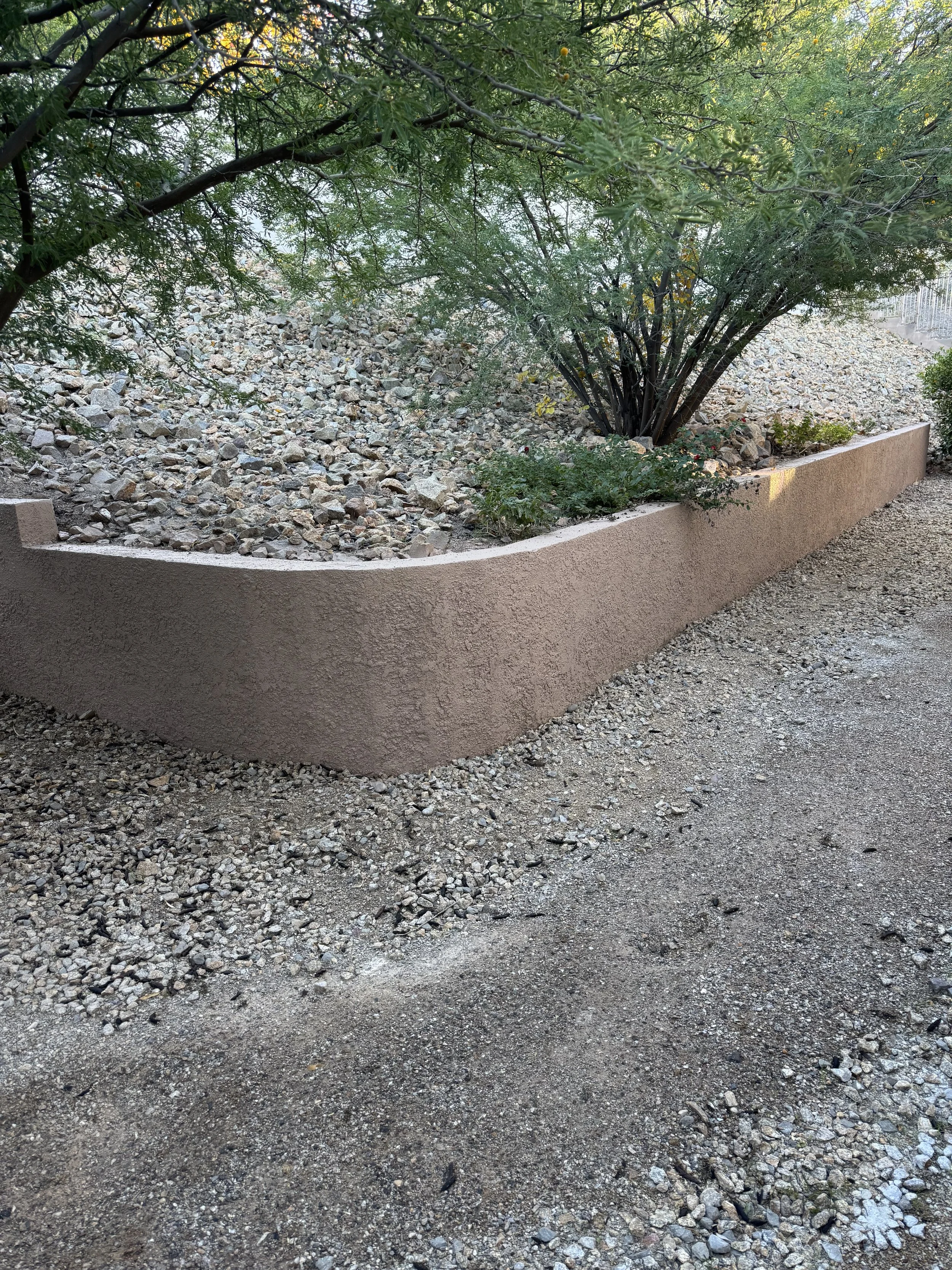A landscaped area with a stucco retaining wall, bushes, and trees with green foliage, surrounded by gravel and dirt ground.