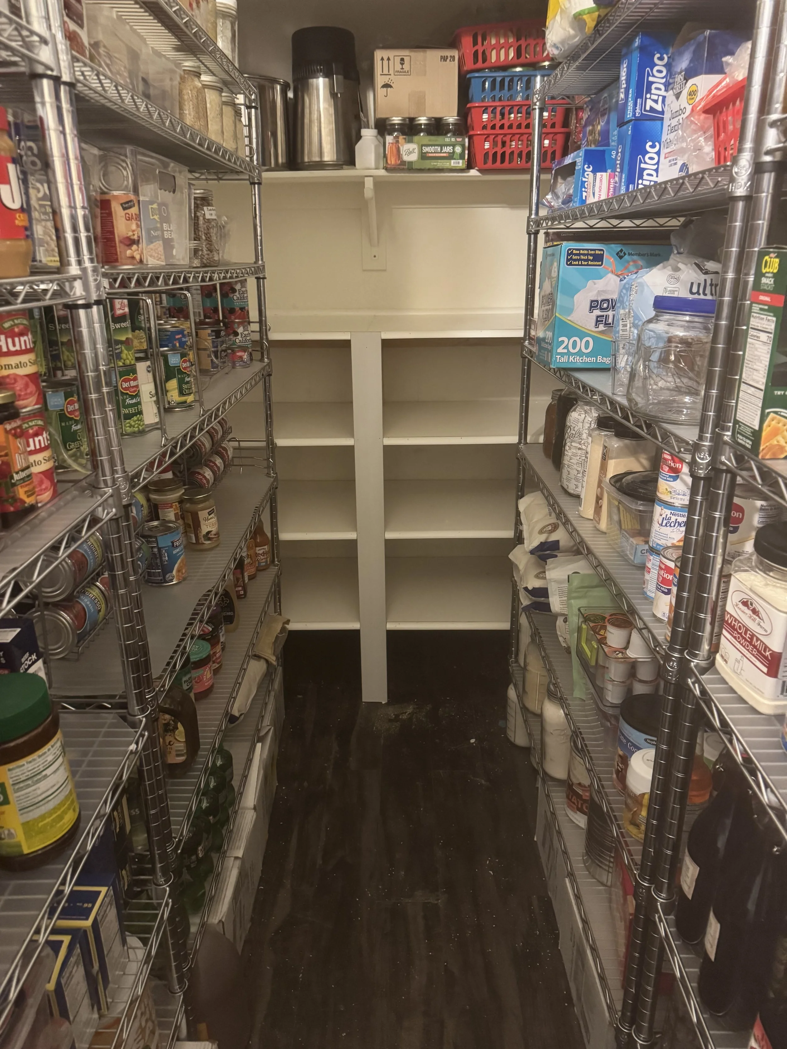 Empty pantry with metal shelves stocked with canned goods, jars, and household items, and a cart at the back.