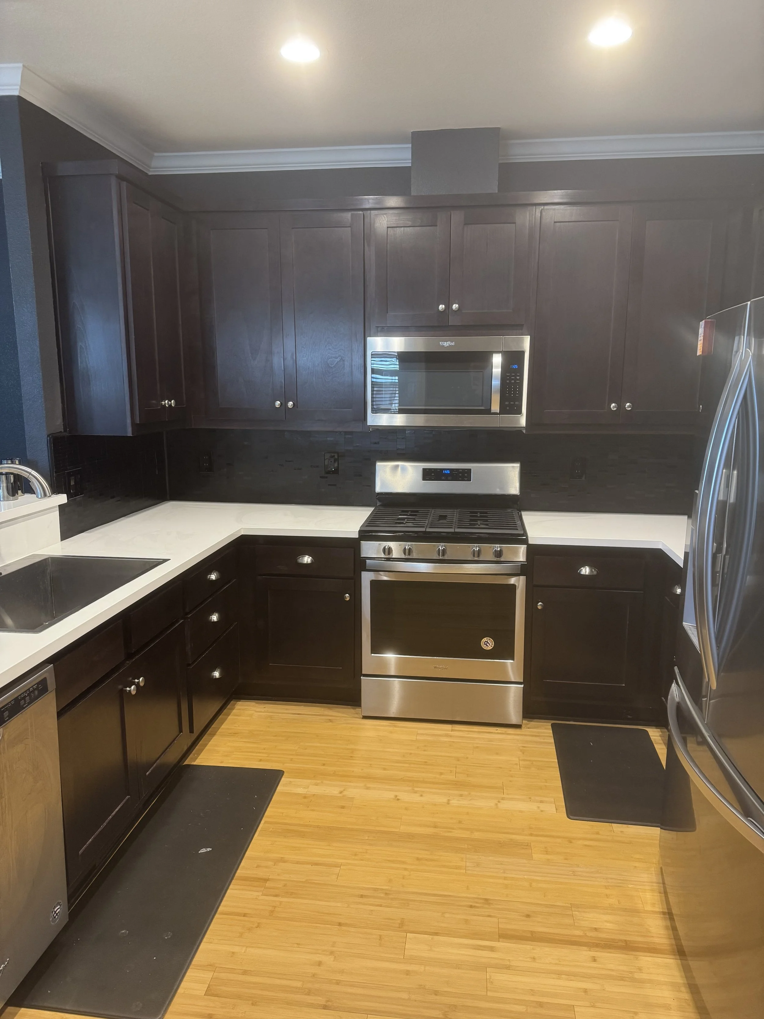 Modern kitchen with dark wood cabinets, stainless steel appliances including microwave and stove, black backsplash, white countertops, and wooden flooring.