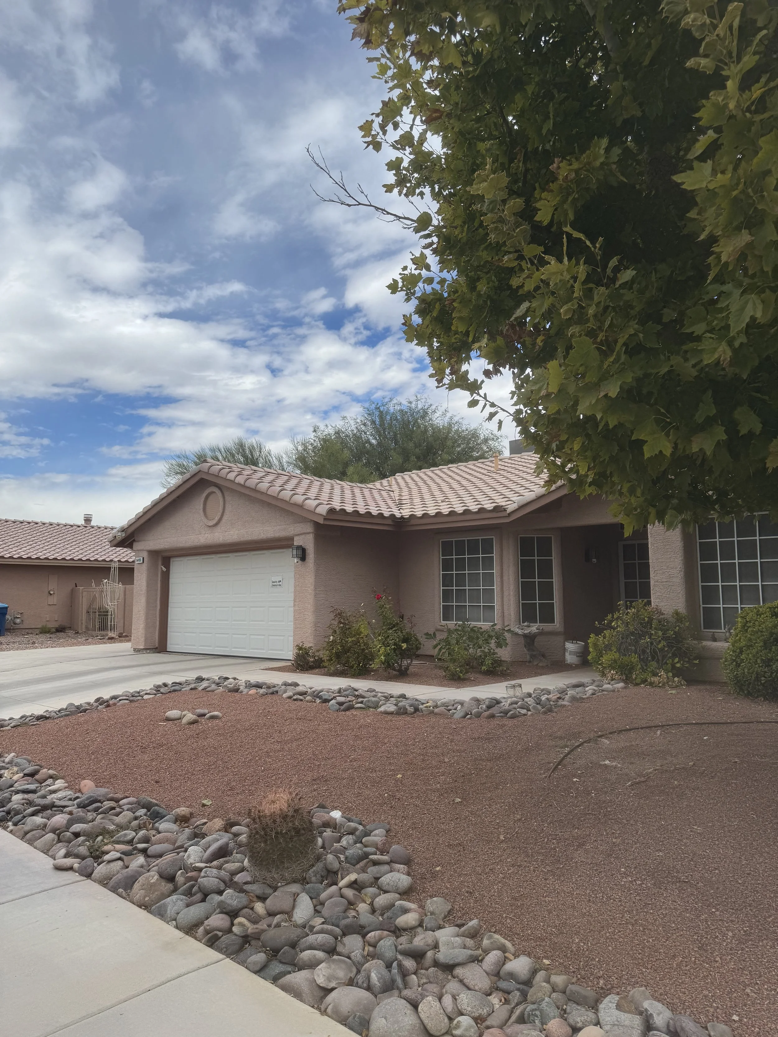 A single-story house with a tiled roof and a two-car garage. The yard features gravel landscaping with rocks and desert plants, and a large tree partially covers the front yard. The sky is partly cloudy.
