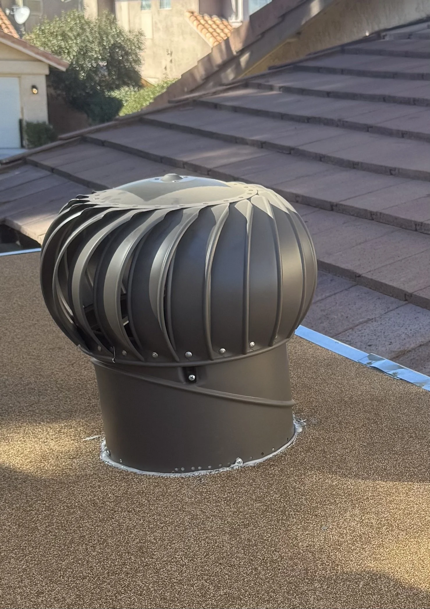 A rooftop with a metallic exhaust vent, surrounded by a pebbled amber color surface, with neighboring rooftops and trees in the background.