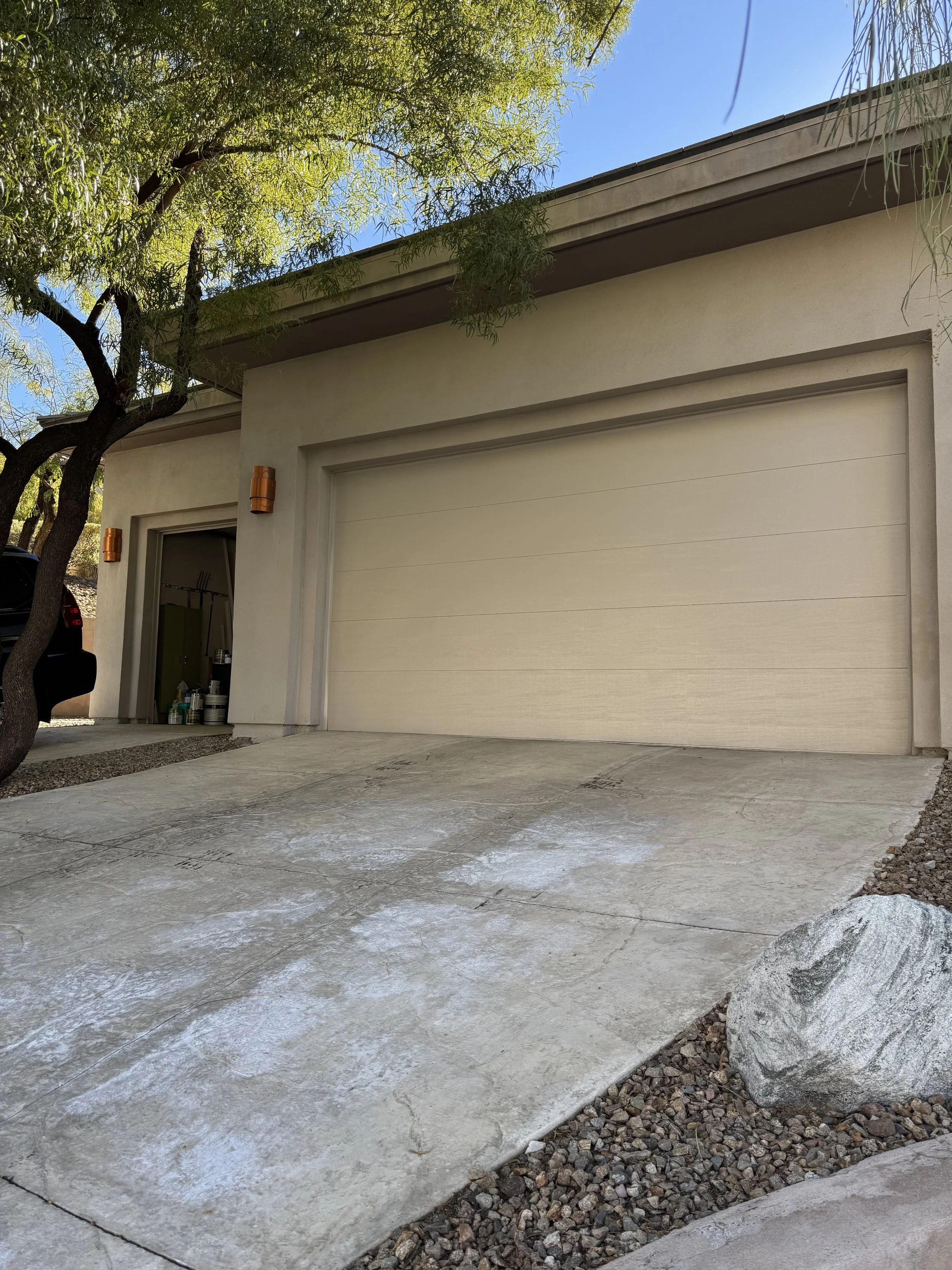 View of a residential house garage with a closed beige garage door, a small open storage area with supplies, a tree with green leaves partially covering the roof, and a gravel and concrete driveway with a large rock.