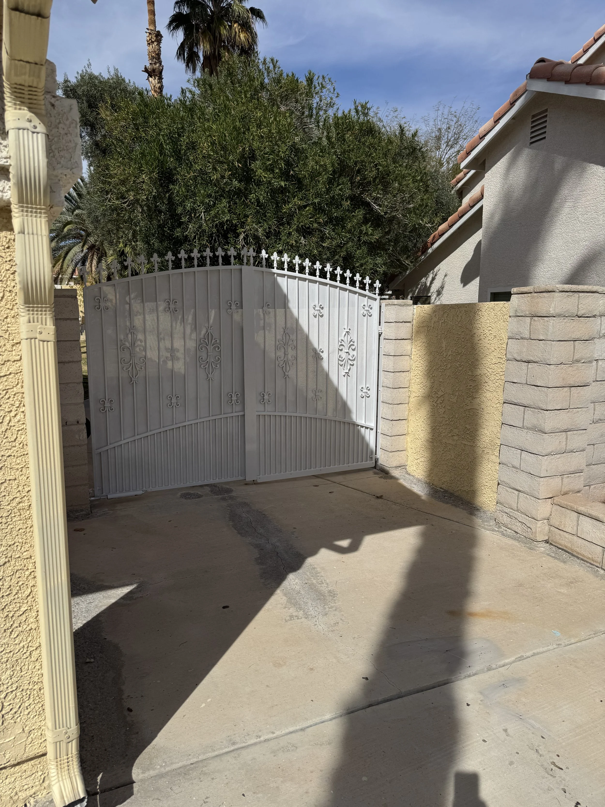 White metal gate with decorative fleur-de-lis patterns, set within a brick and stucco wall, casting shadow on concrete driveway with a backdrop of tall trees and a partly cloudy sky.