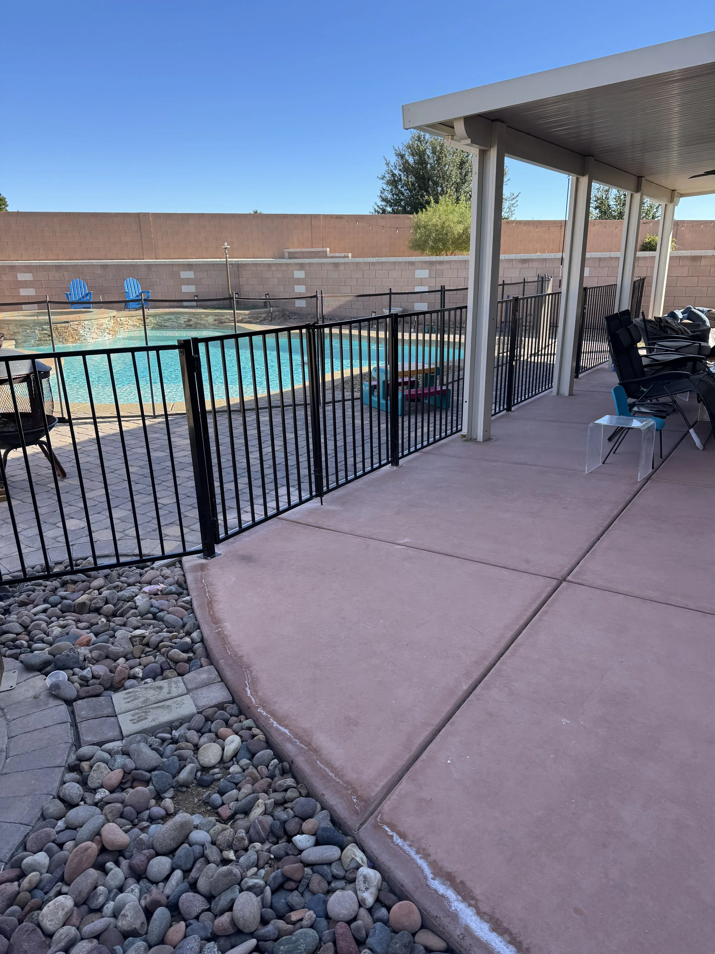 View of a backyard swimming pool with a black safety fence, surrounded by a concrete patio and rocks, with pool furniture and two blue chairs in the background under a clear sky.