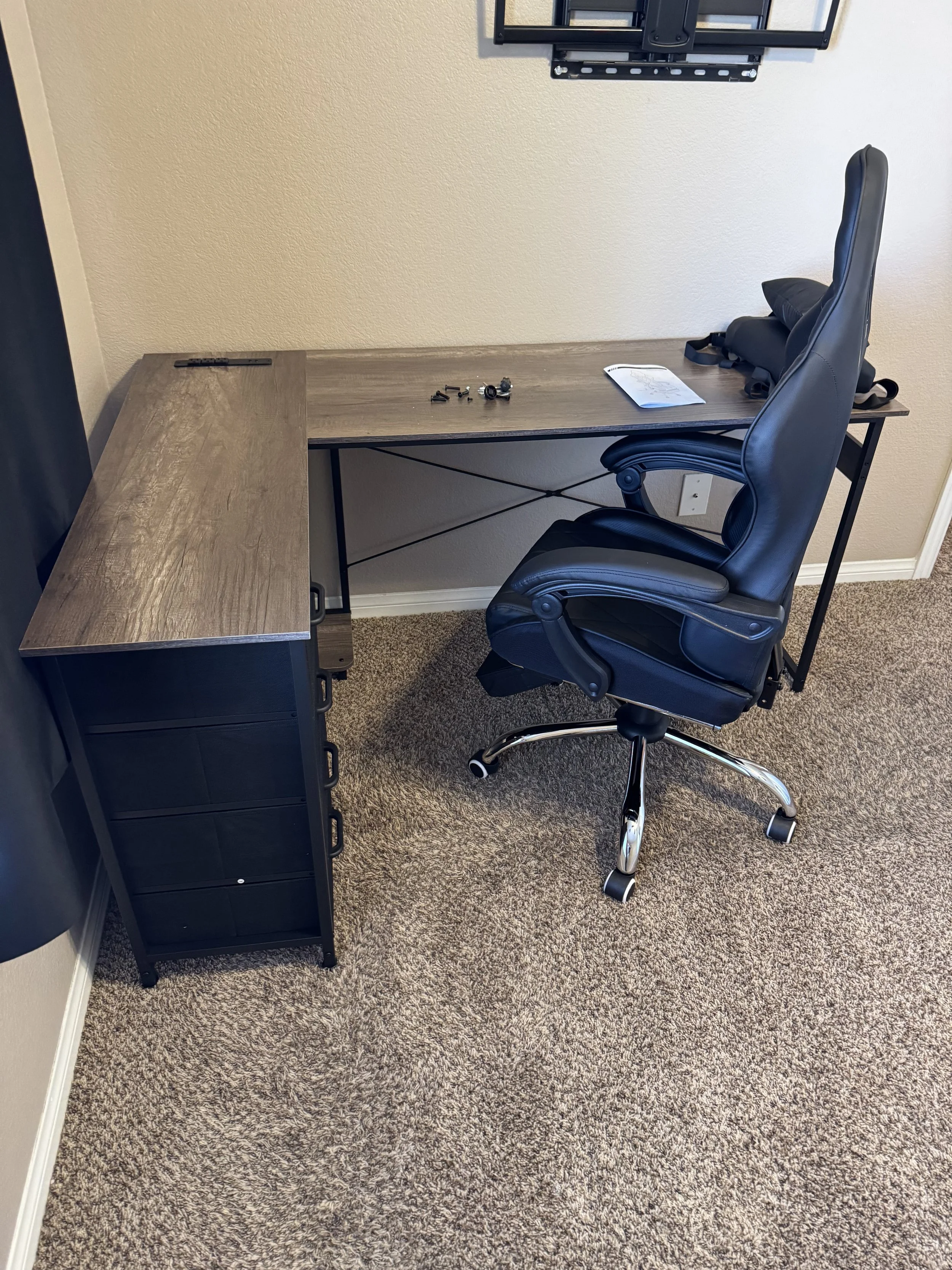 Empty office desk with a black ergonomic chair, some screws, and a paper on the desk, with a wall-mounted TV above.