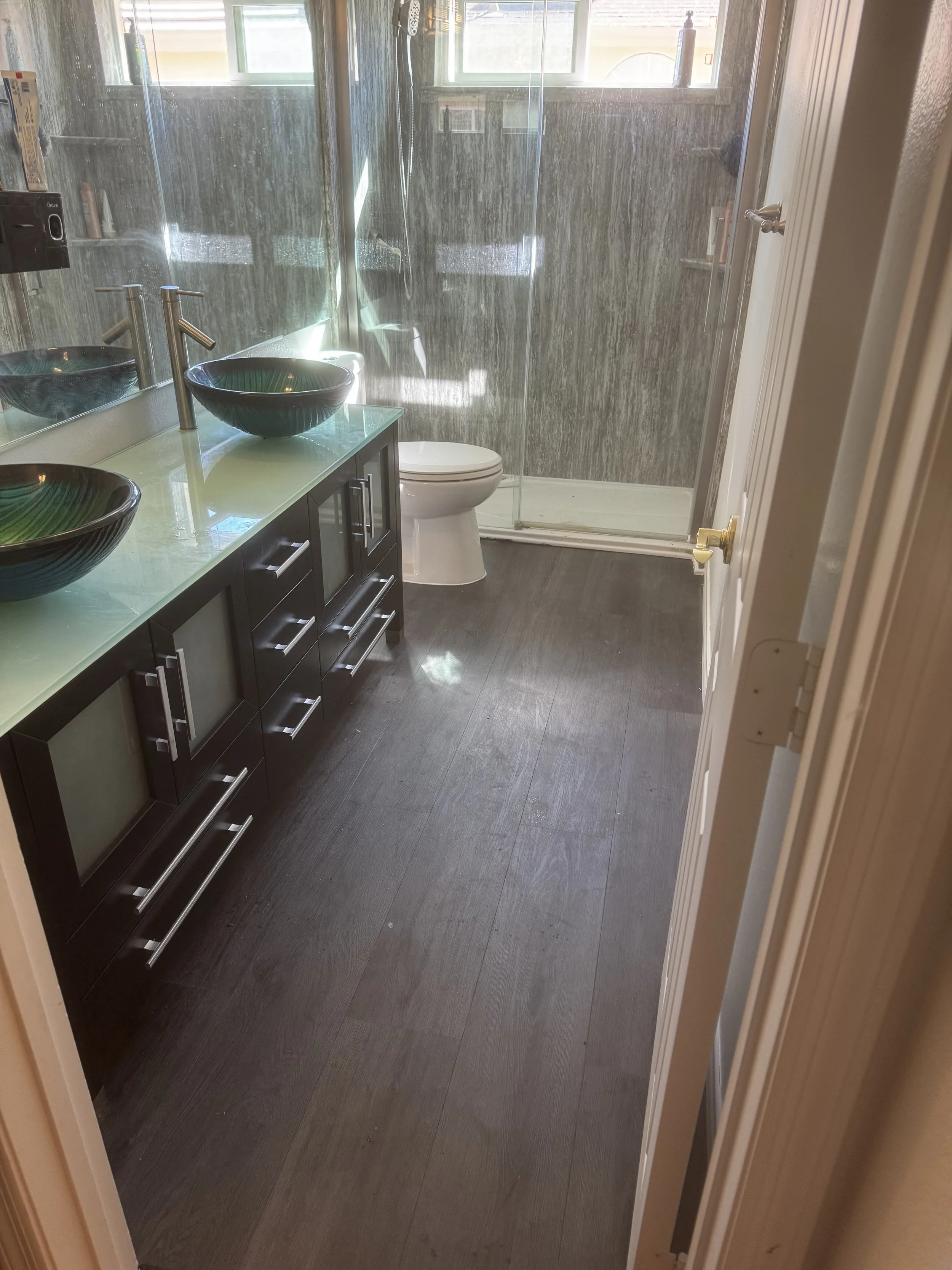 Bathroom with two black vessel sinks with green interiors, a large mirror, a dual-sink vanity, a toilet, and a glass-enclosed shower with tiled walls, wooden flooring, and natural light from windows.