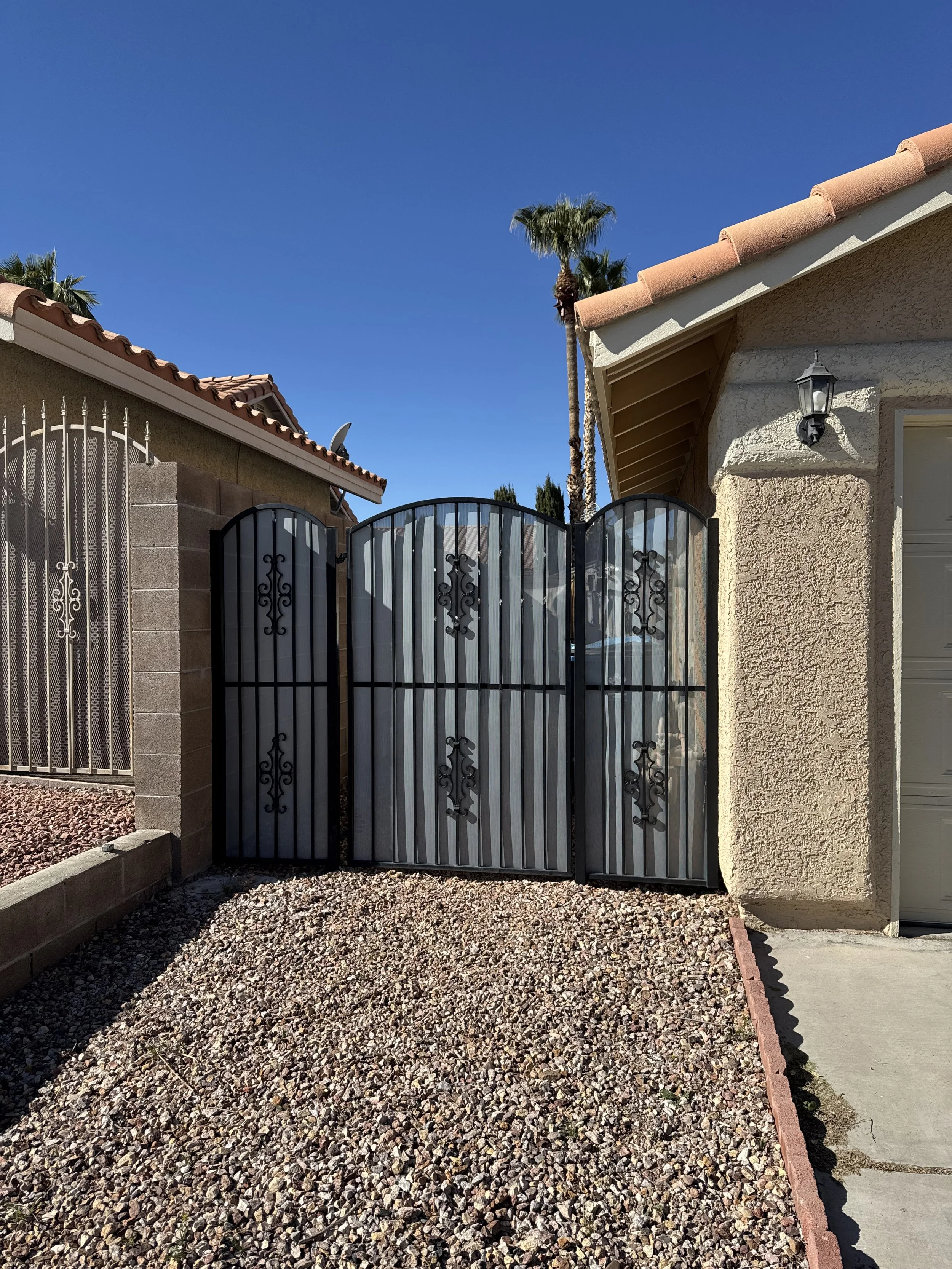Decorative black metal gate with scrollwork design, located between two houses with desert landscaping and a clear blue sky.