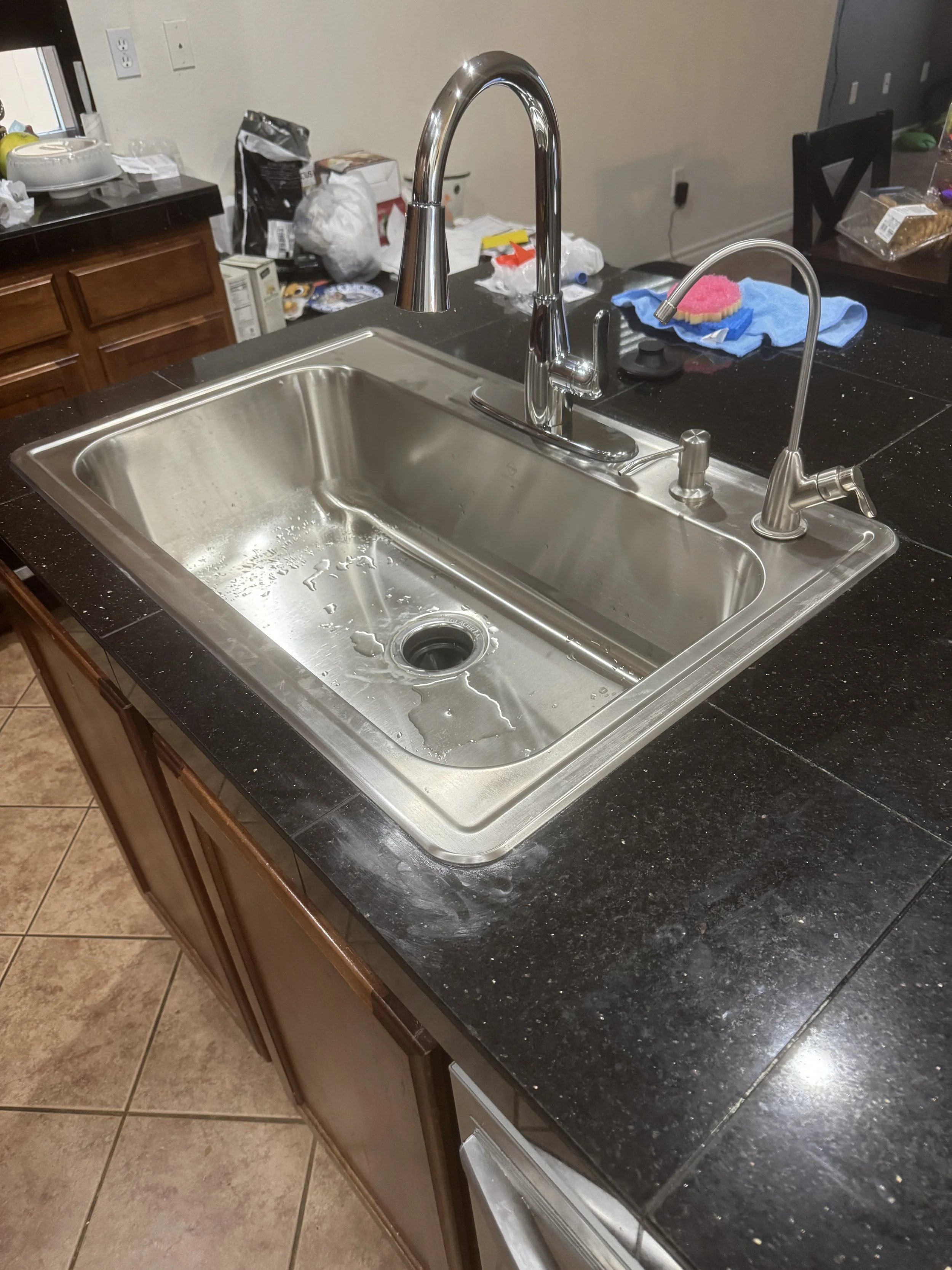 Stainless steel kitchen sink with water droplets, installed in a black countertop, with a high-arc faucet and a soap dispenser. Kitchen clutter including cleaning supplies and dishes are visible in the background.